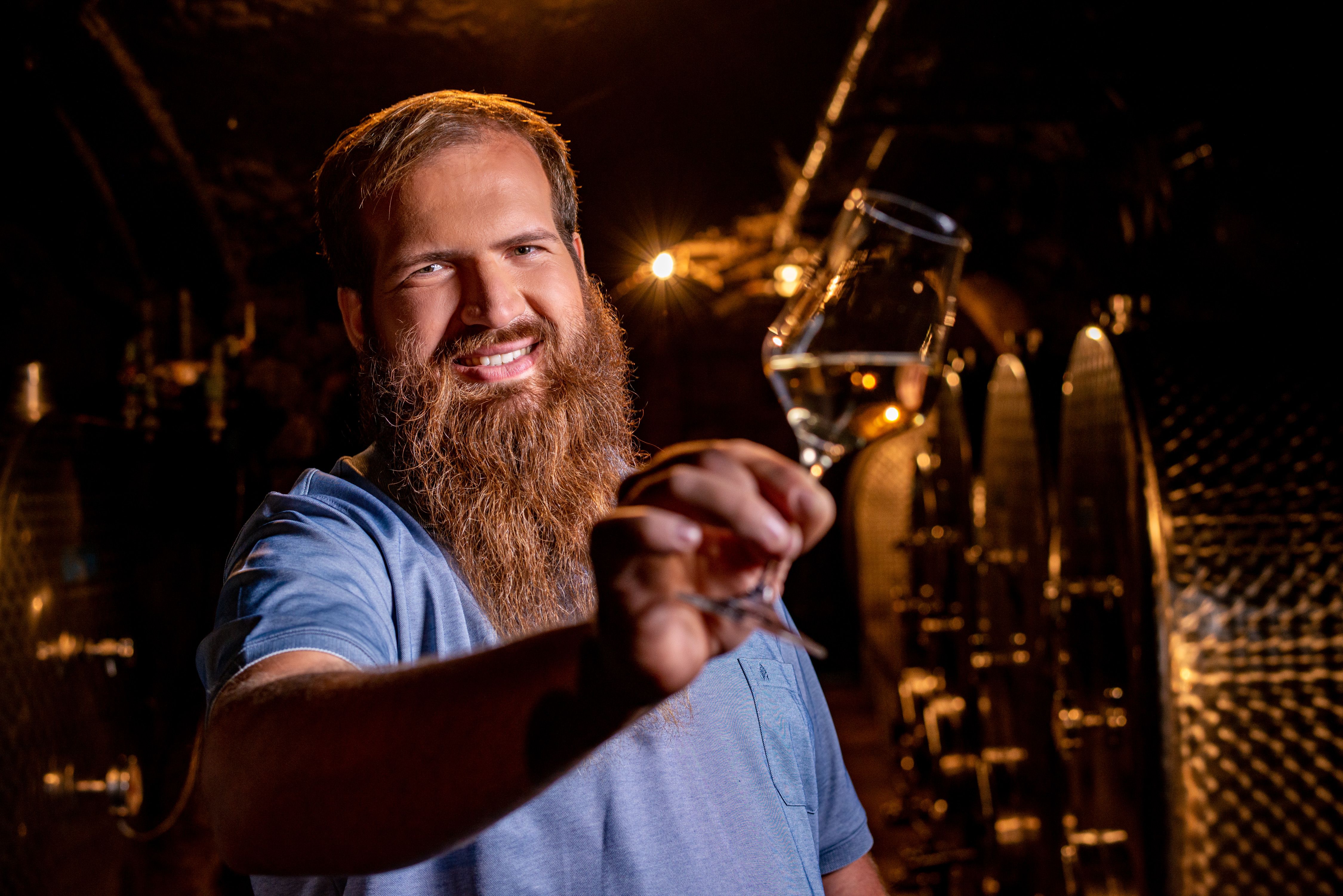 Man with beard holding a wine glass in a wine cellar.