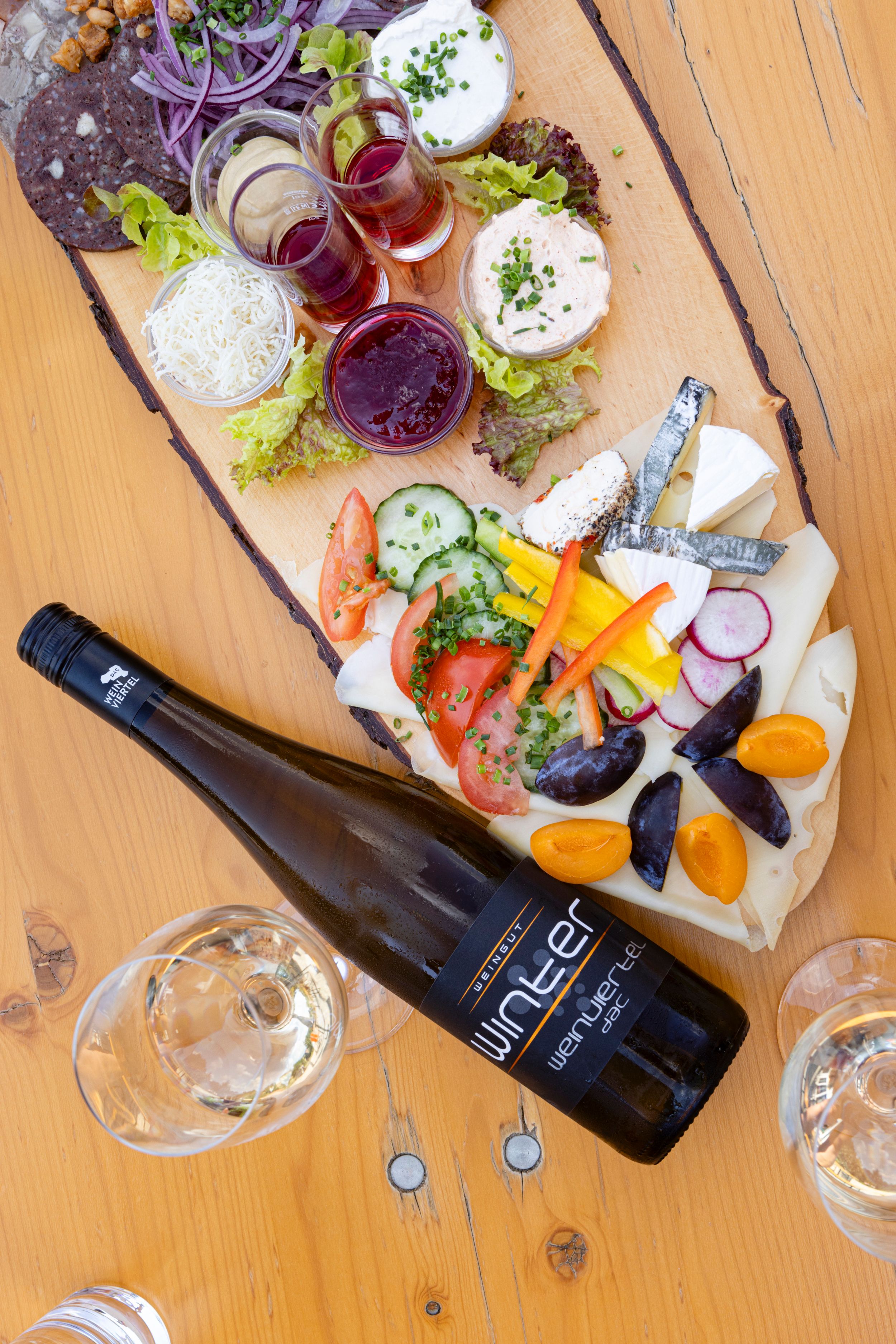 A Heurigen platter with various cheeses, spreads, vegetables and a bottle of wine on a wooden table.