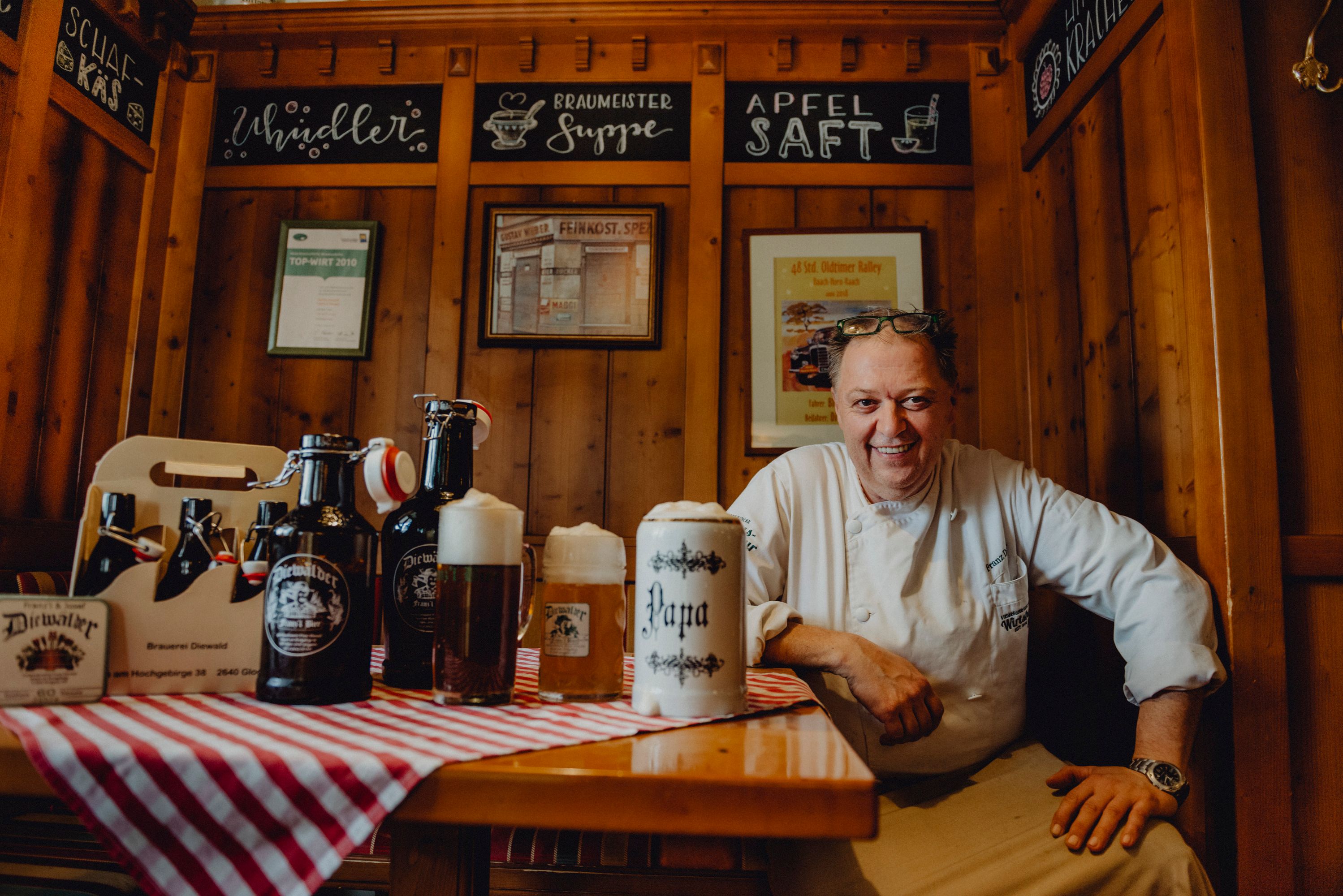 A landlord in traditional dress sits smiling at a table with beer mugs and bottles in a rustic dining room.