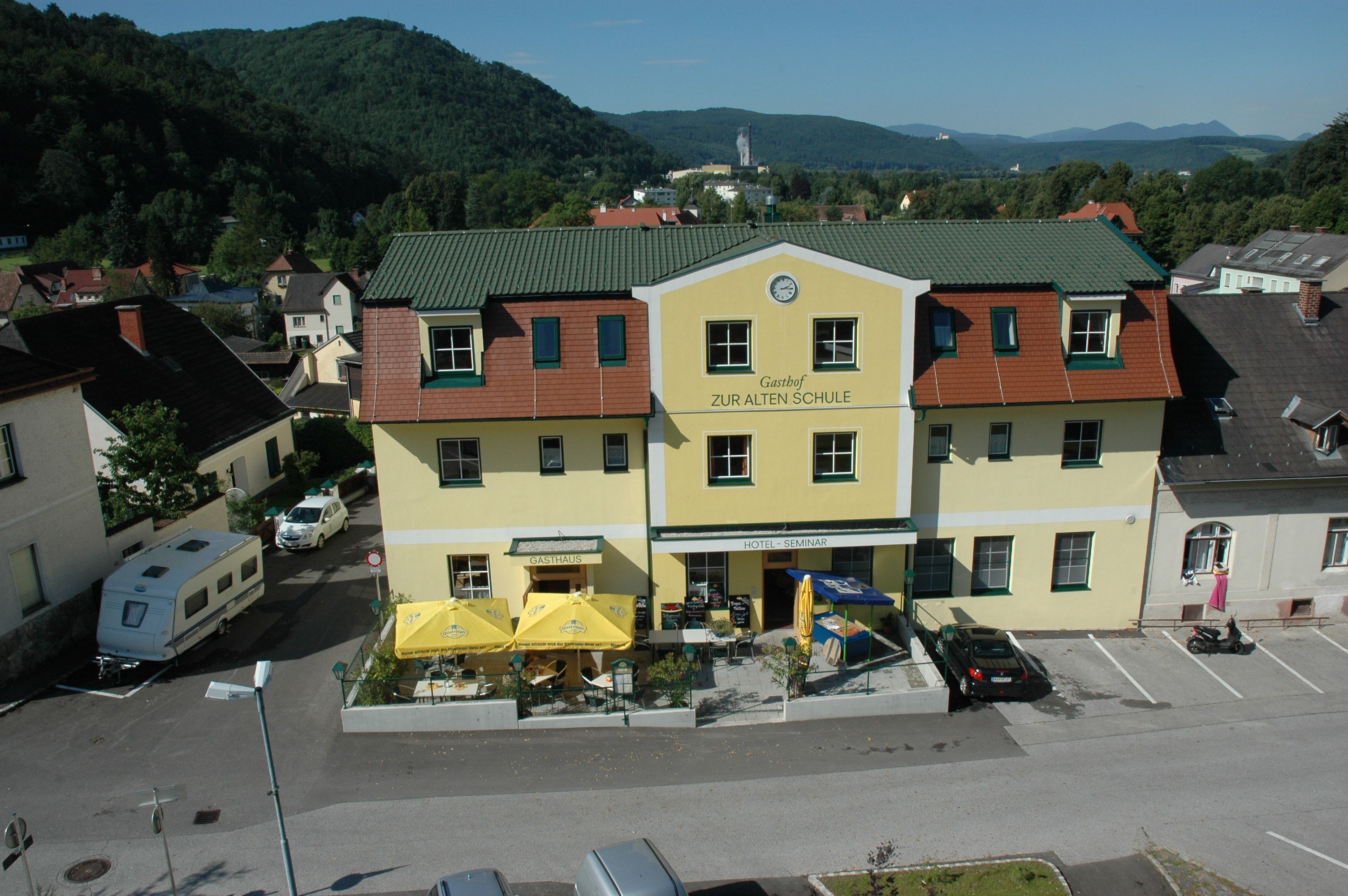 Yellow building with the inscription 'Gasthof Zur Alten Schule', surrounded by hills and trees.