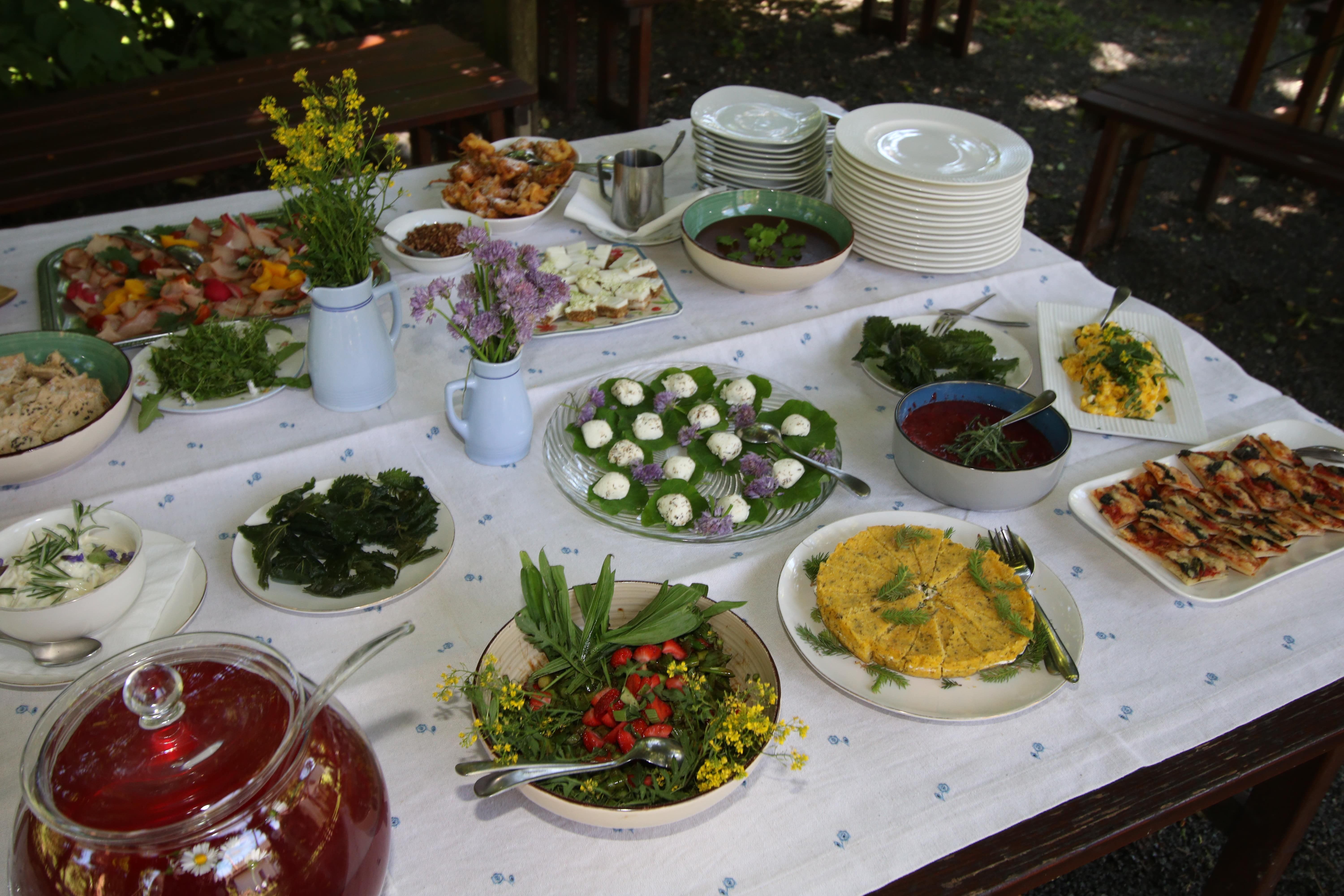 Table with white tablecloth and various dishes such as salads and cheese, next to plates and flower vases.