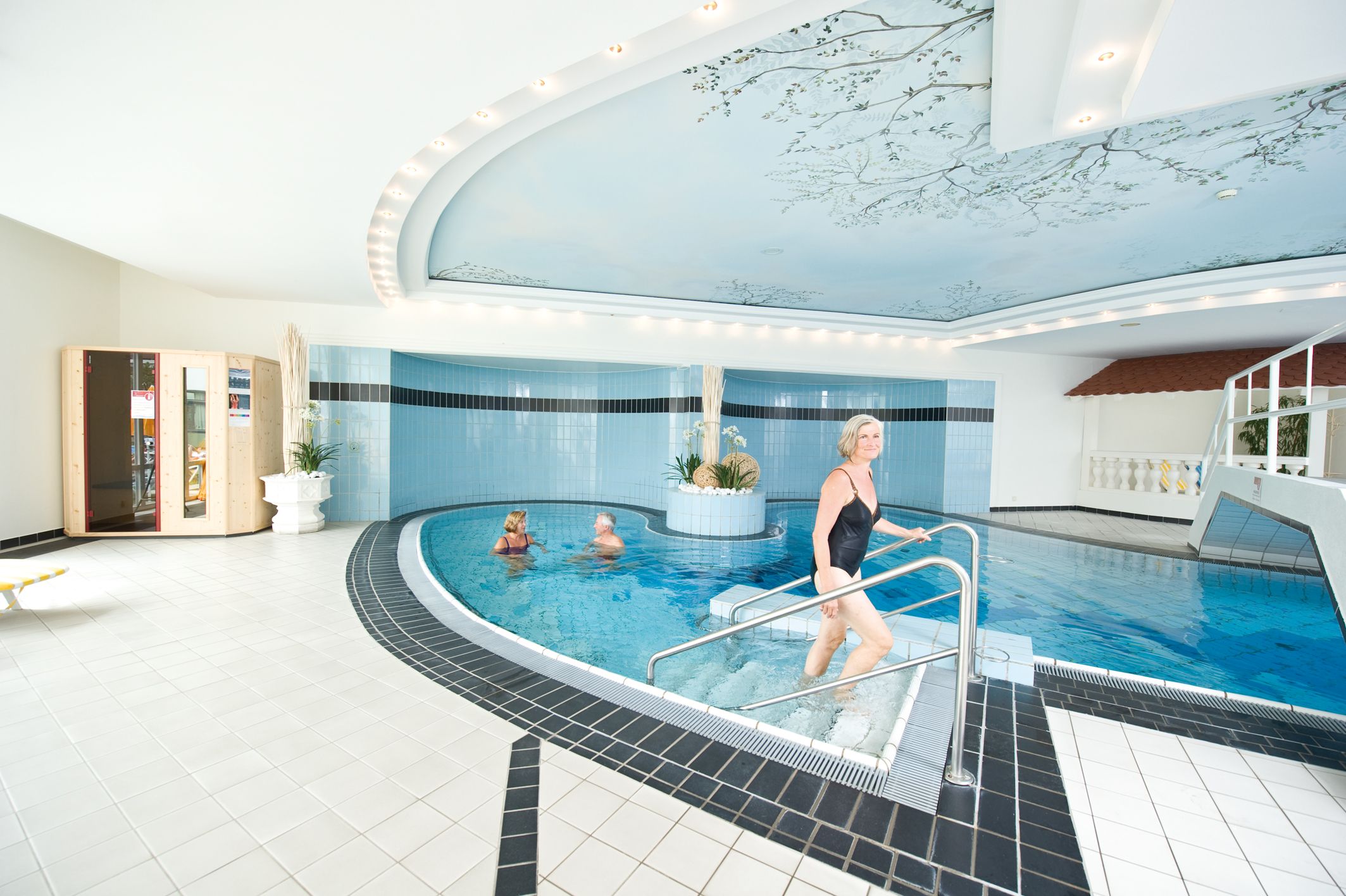 Indoor pool with people in the water and a woman walking up the stairs.