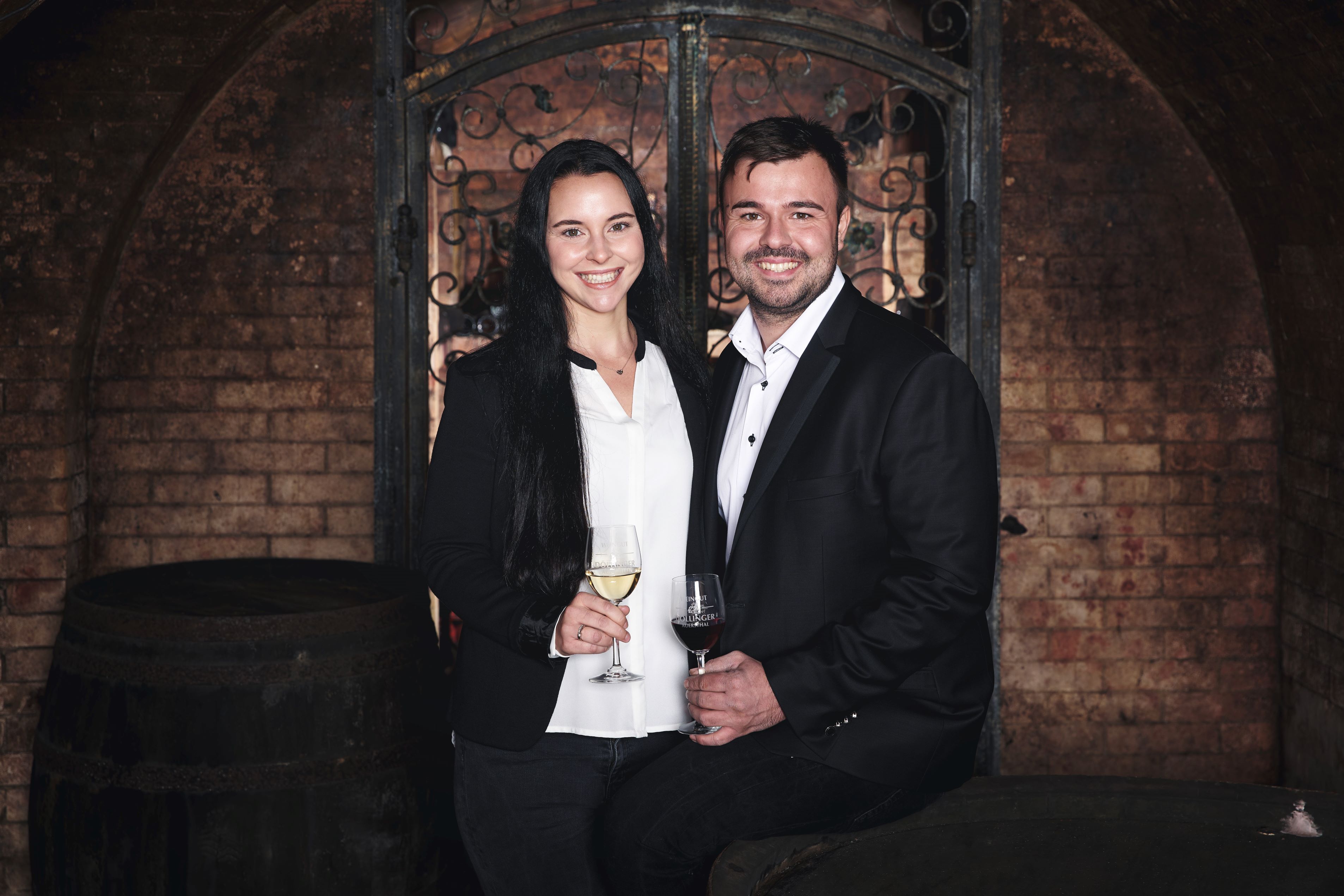 A man and a woman in elegant clothes are standing in a wine cellar with wine glasses in their hands.