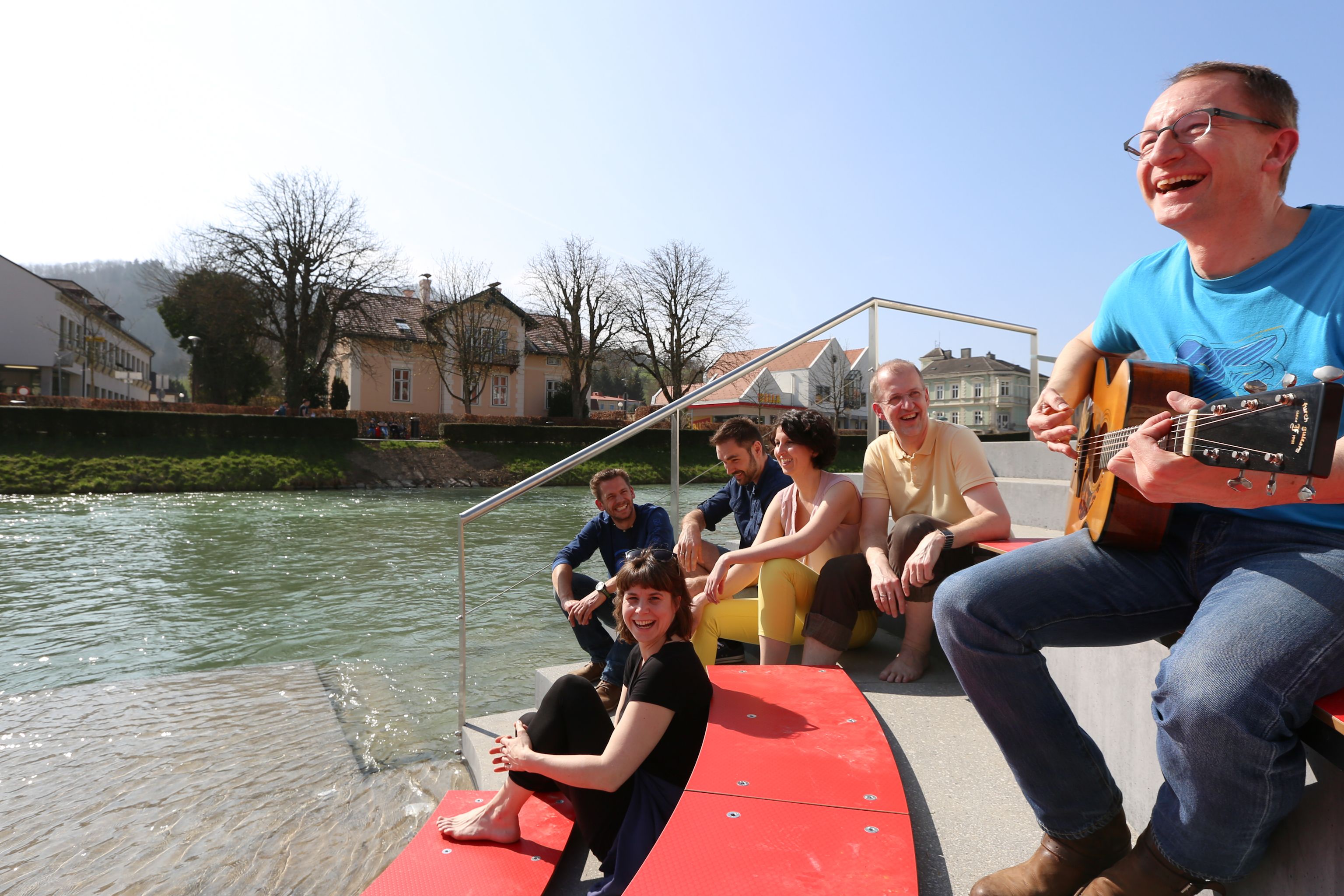 Group of people sitting on a riverbank laughing, one person playing the guitar.