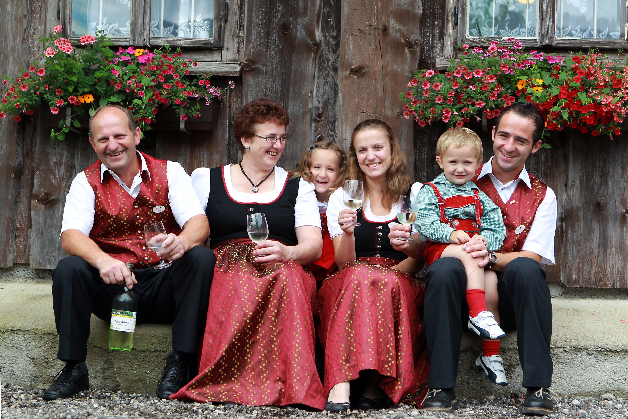A family in traditional dress sits in front of a wooden house with flowers and holds glasses of wine.