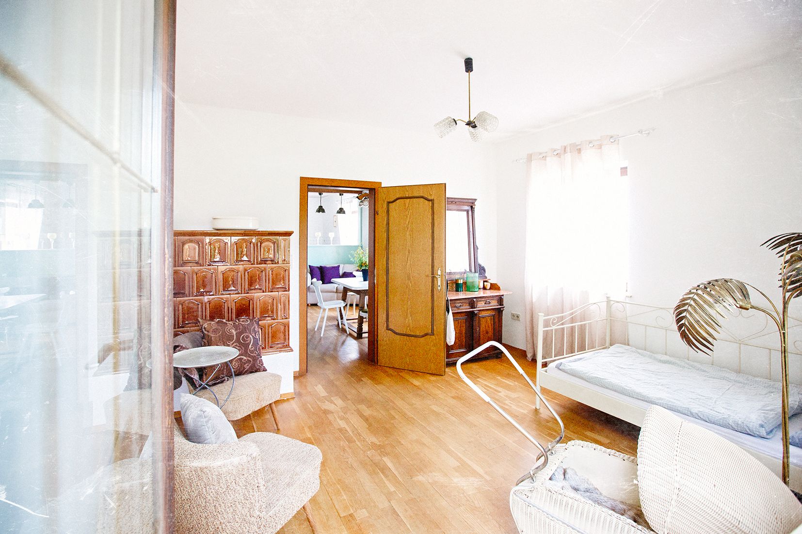 A bright room with a wooden floor, a bed, an armchair, and a passageway leading to another room with a dining table.