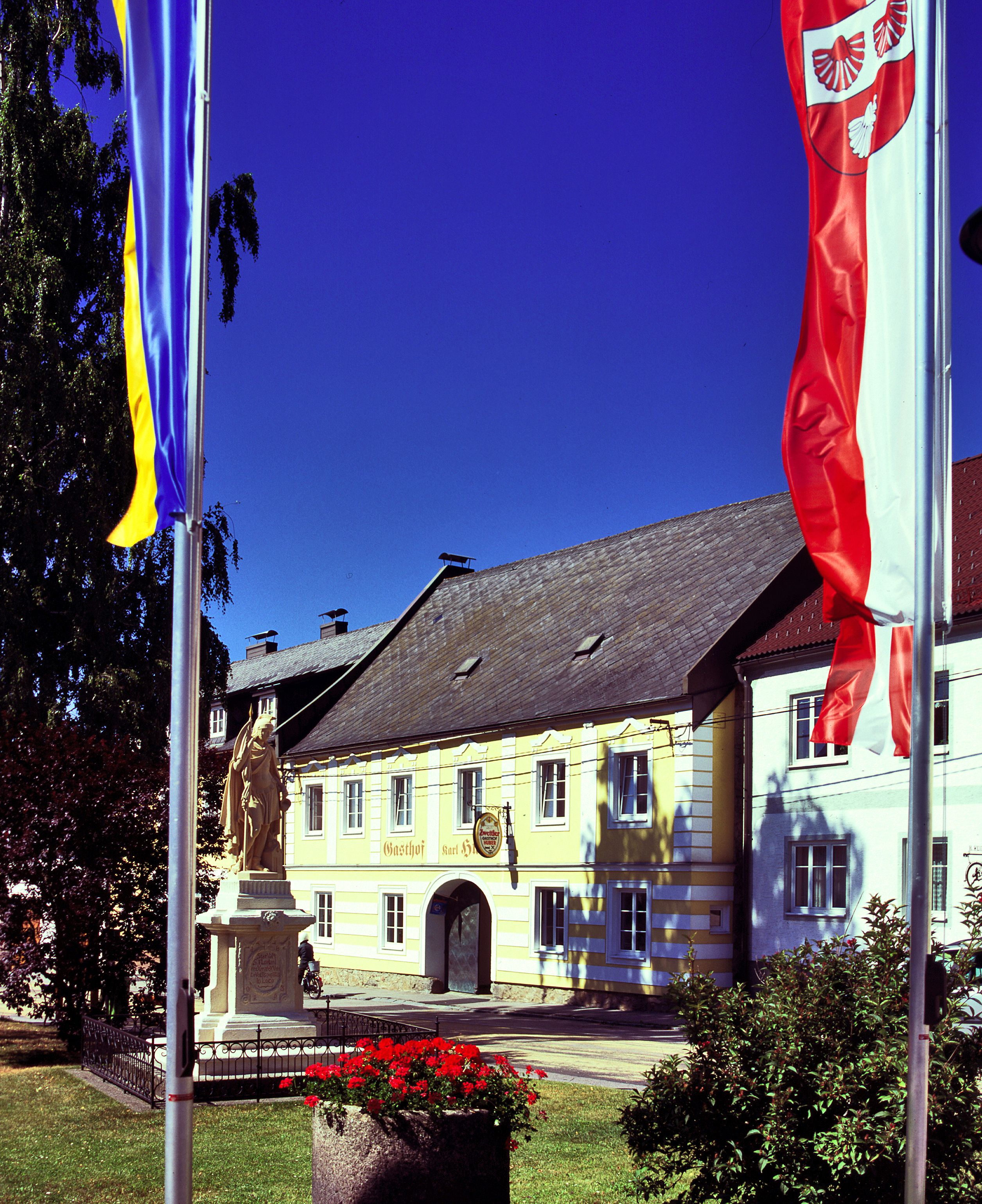 Yellow building with the inscription 'Gasthof Karl Huber', surrounded by flags and a statue.