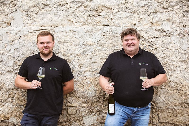 Two men in black polo shirts with wine glasses in front of a stone wall.