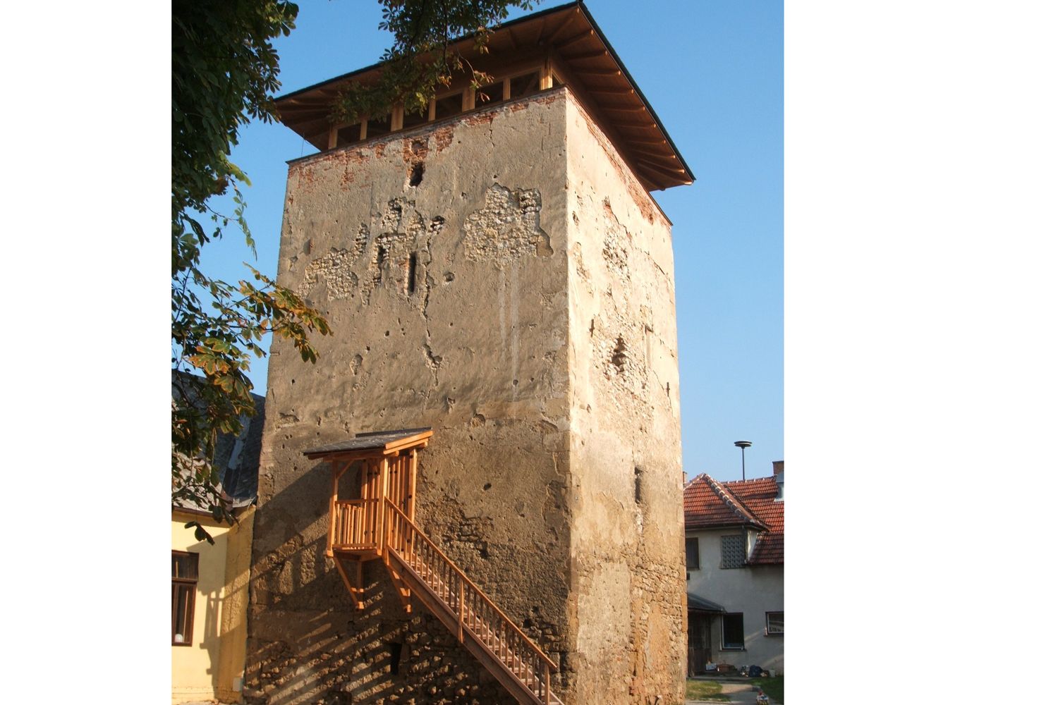 An old fortified tower with a crumbling façade and a wooden staircase, surrounded by houses and trees.
