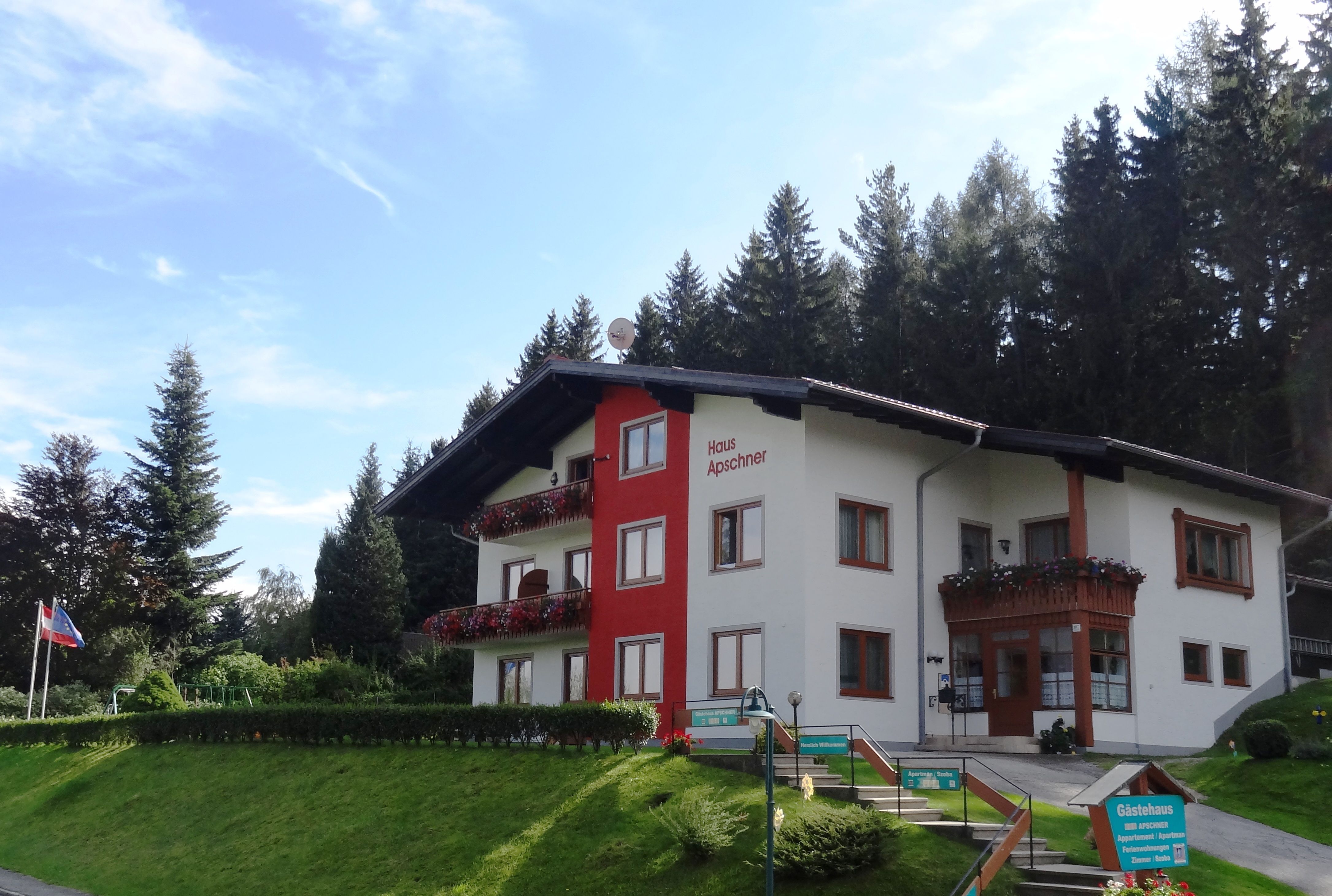 A two-storey guest house with red balconies and flowers, surrounded by trees and a blue sky.