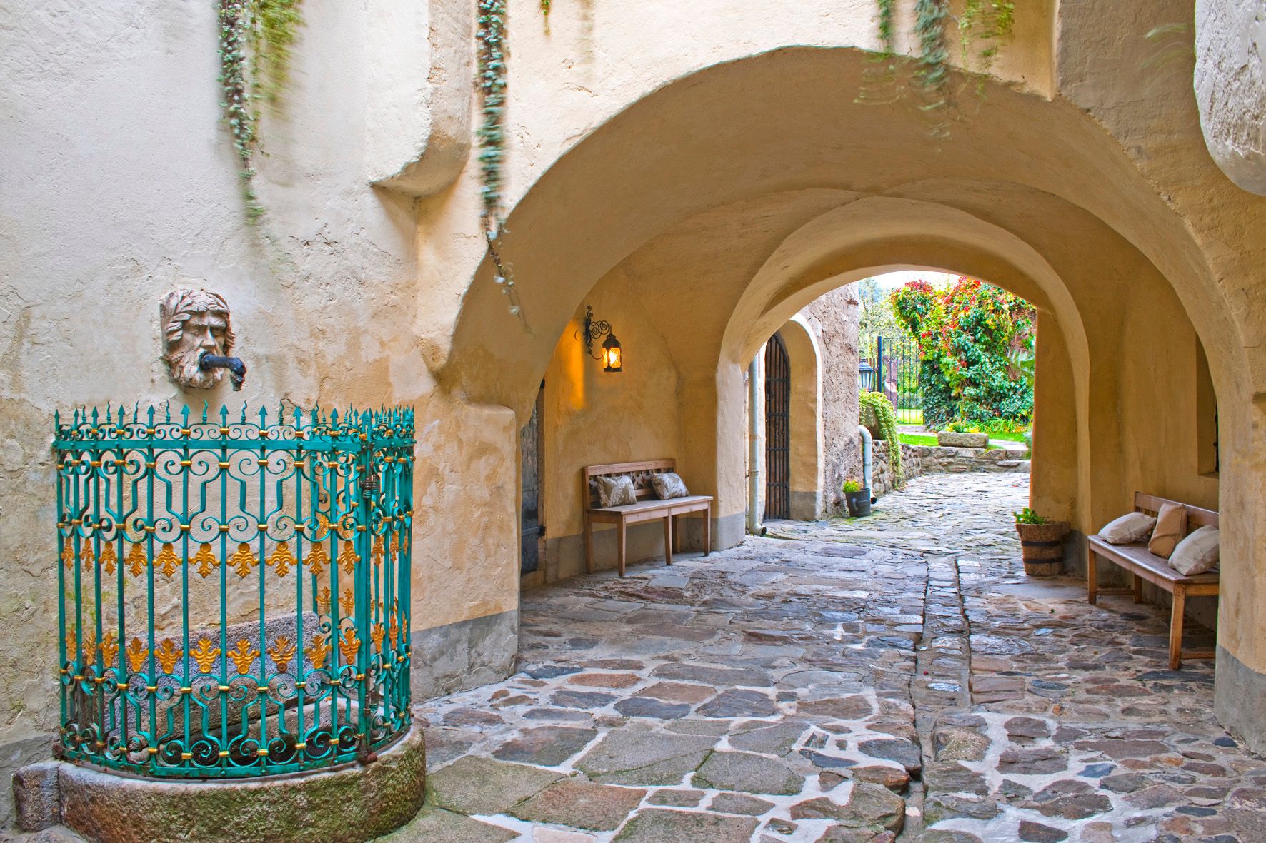 A stone passageway with benches, a lantern and an ornate fountain.