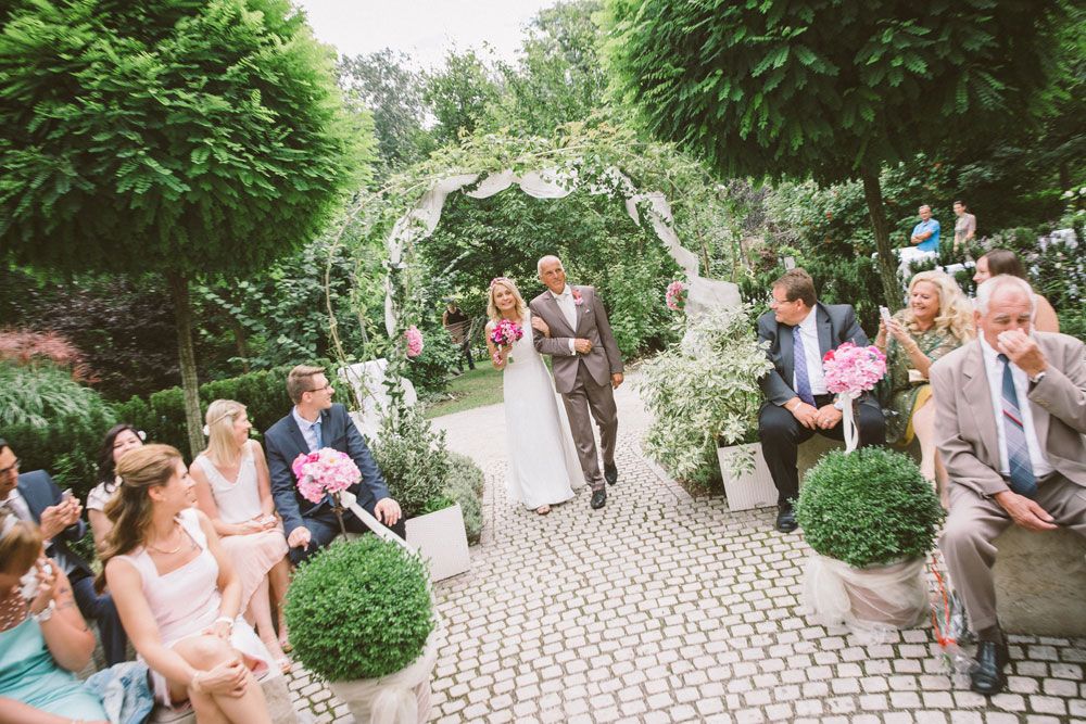Bride and attendant walk through a garden arch, surrounded by guests.