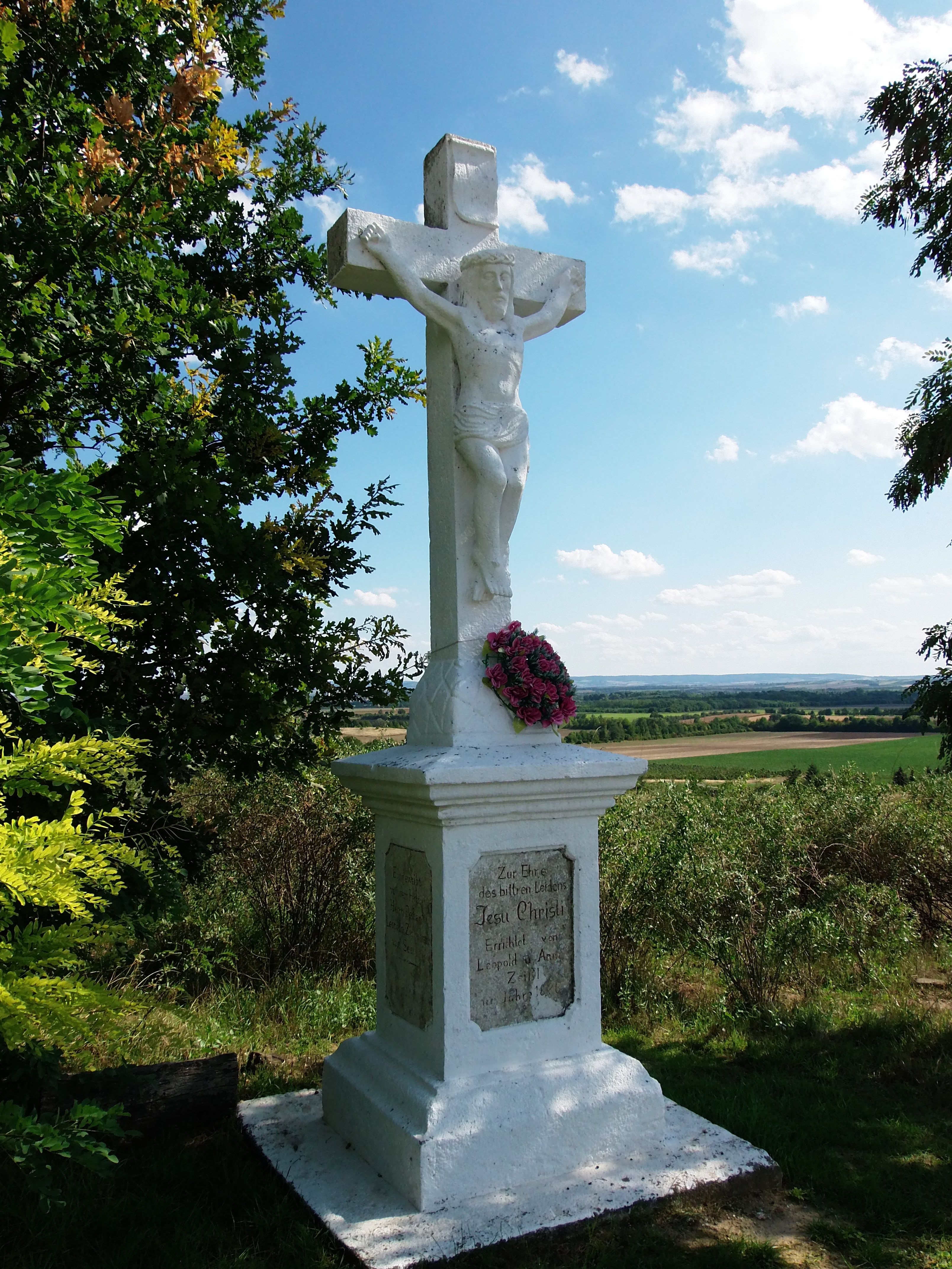 White stone cross with figure of Jesus and wreath of flowers, surrounded by trees and landscape in the background.