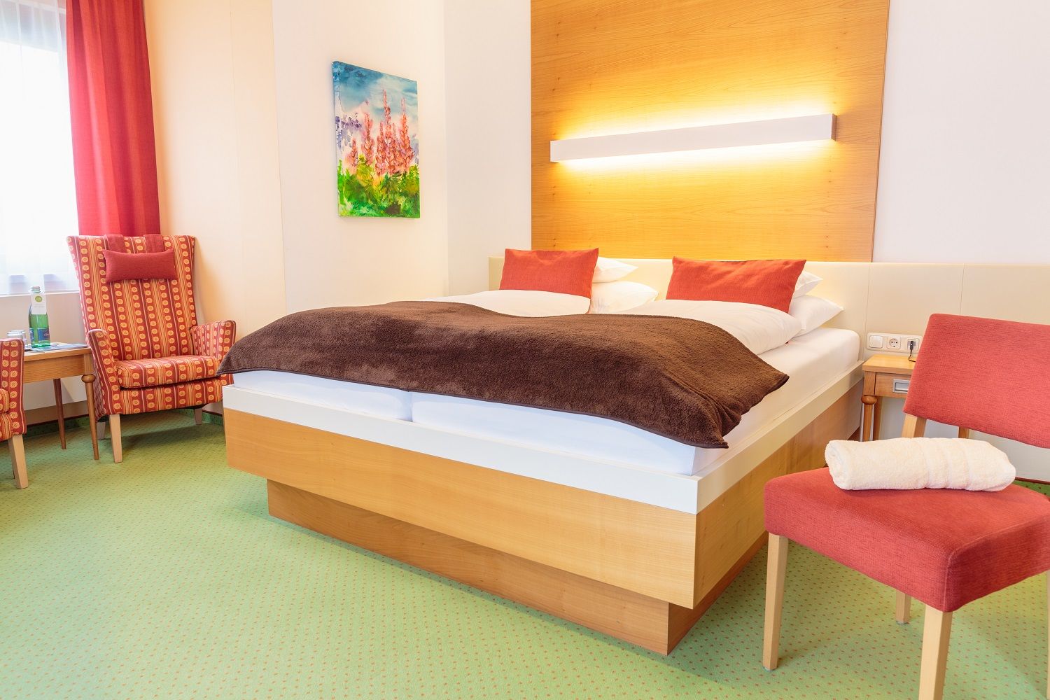 A cozy double room with a large bed, red cushions, an armchair, a chair and a painting on the wall.