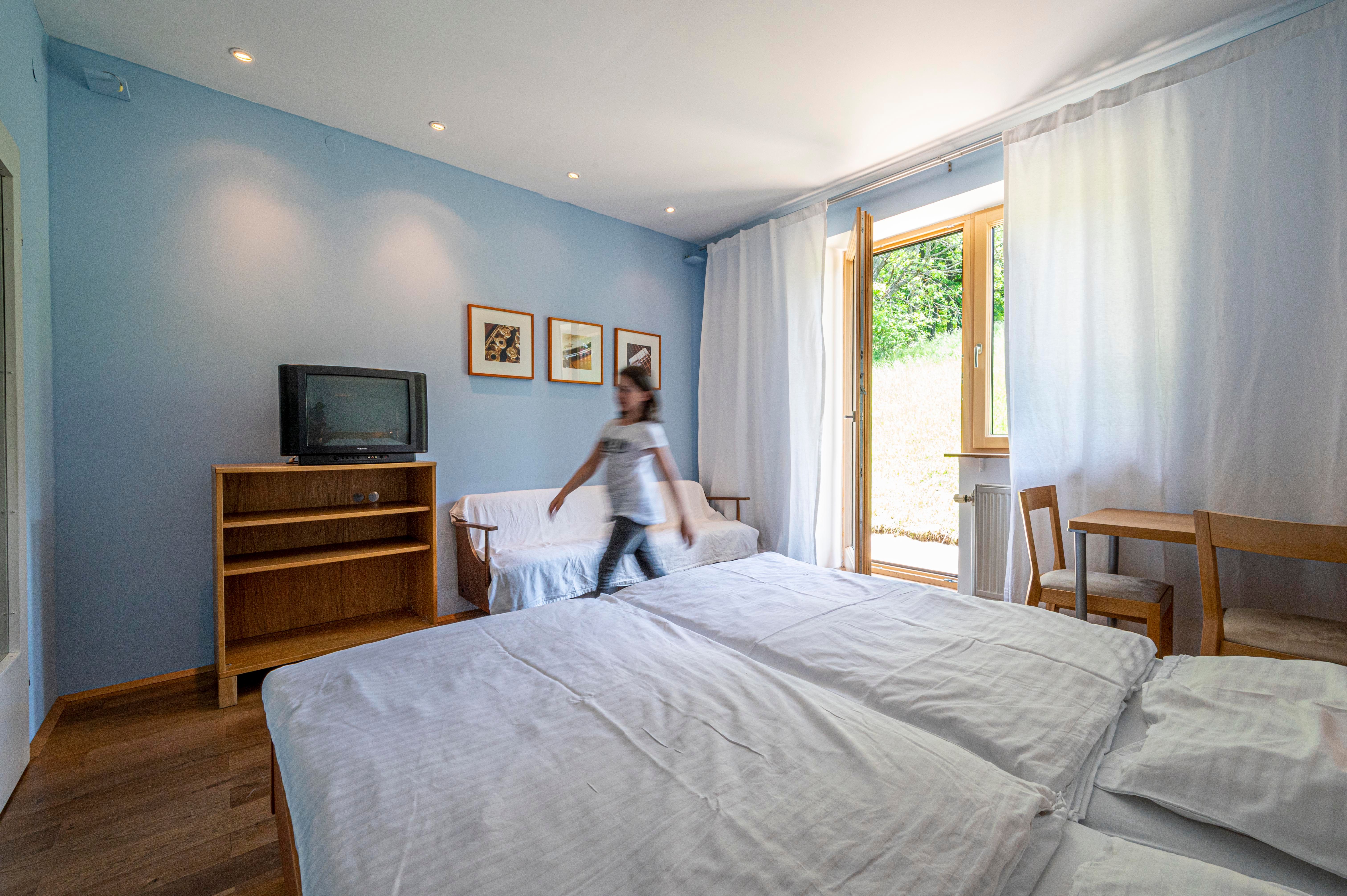 A bright room with blue walls, a bed, a television and a small table. A person walks through the room.