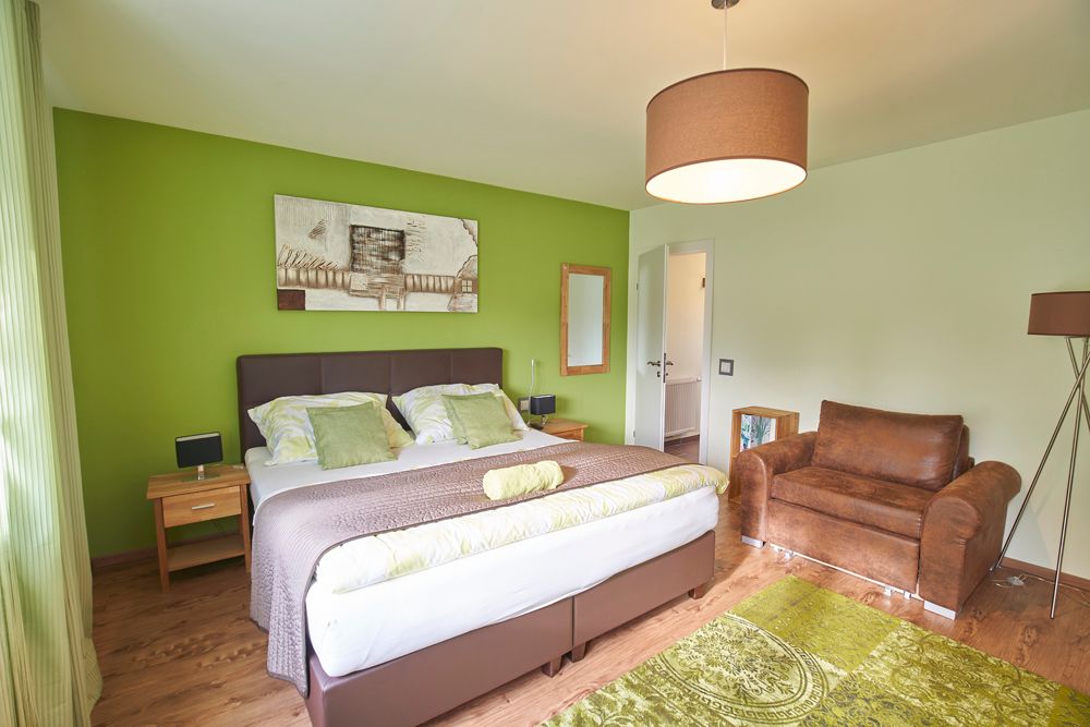 Modern furnished guest room with green accent, double bed, brown armchair and decorative picture on the wall.
