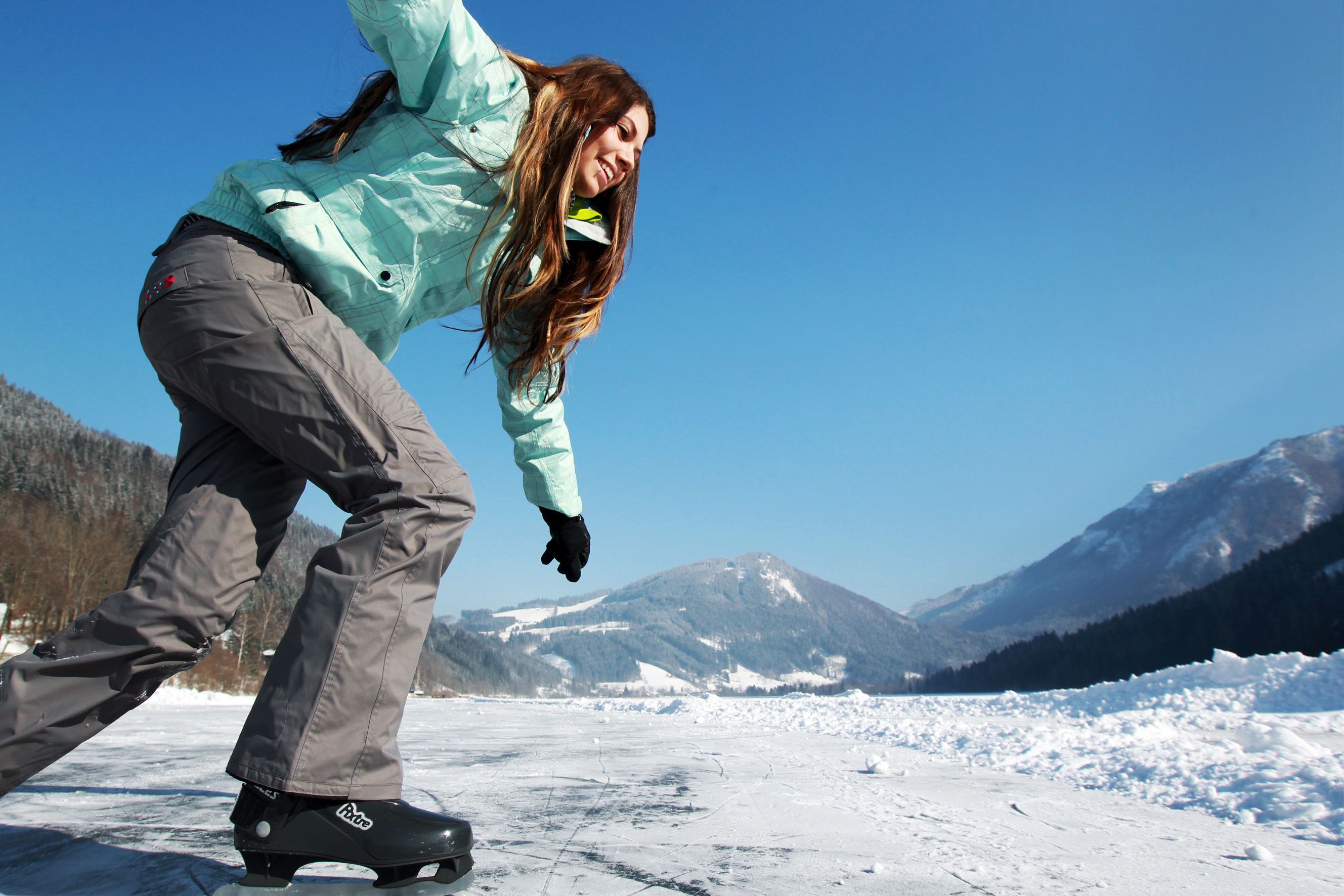 A person skates on a frozen lake with snow-covered mountains in the background.