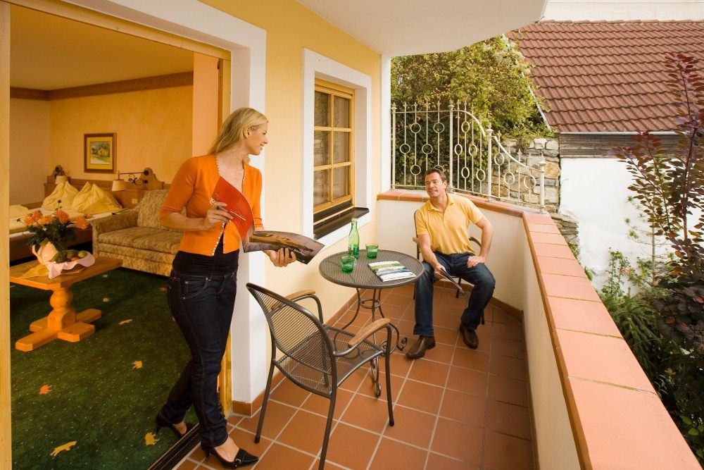 A man sits on a balcony, a woman comes out of the room with a magazine. Table with drinks, cozy atmosphere.