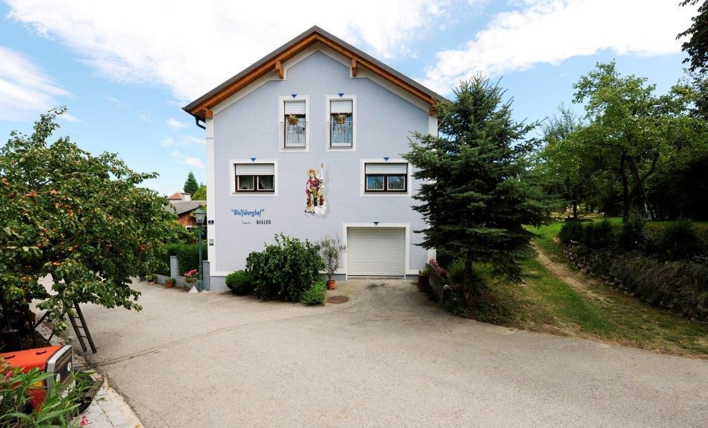 A two-storey house with a blue façade and wooden roof, surrounded by trees and a paved path.