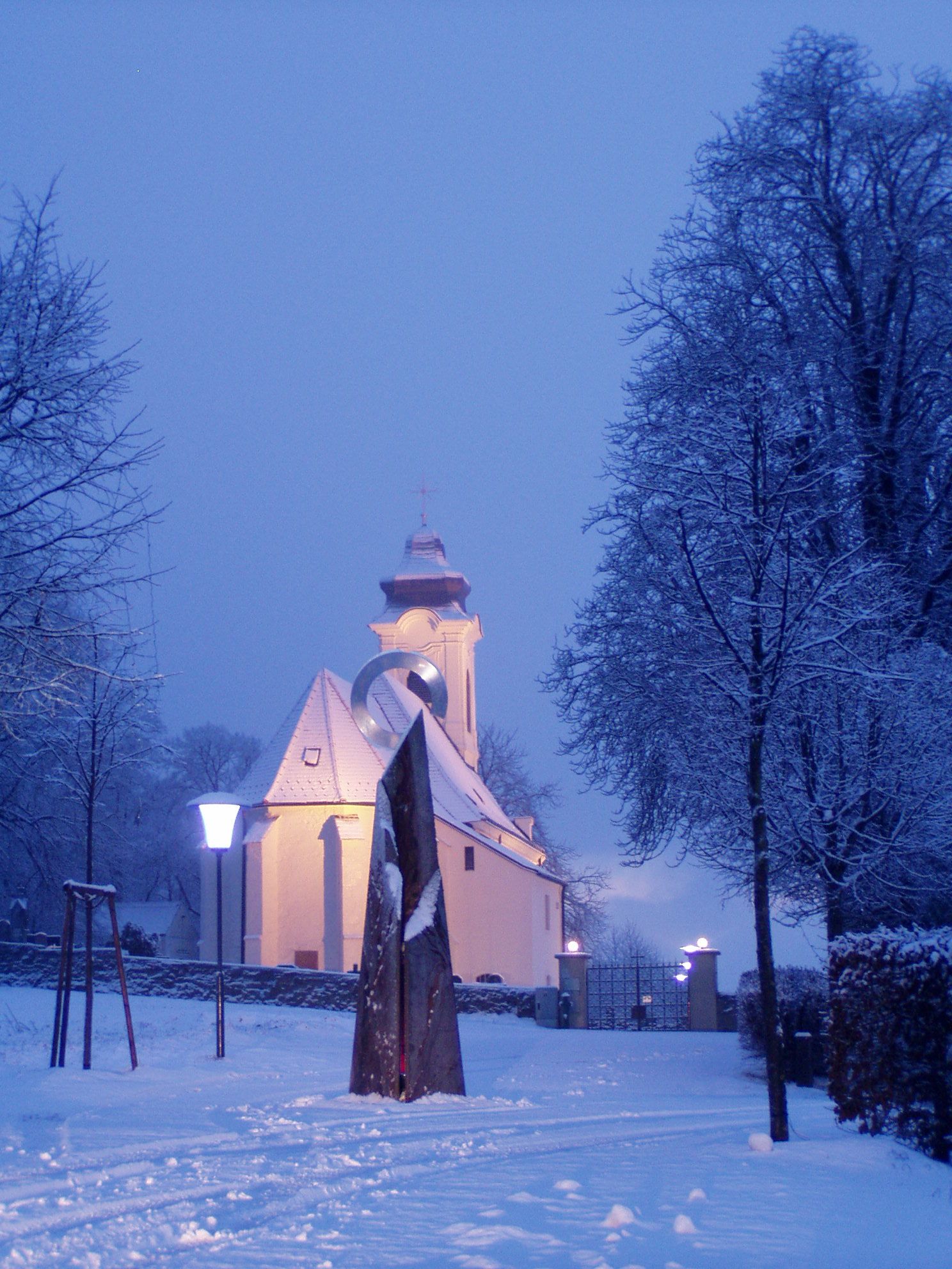 A snow-covered church with an illuminated tower and a sculpture in the foreground.
