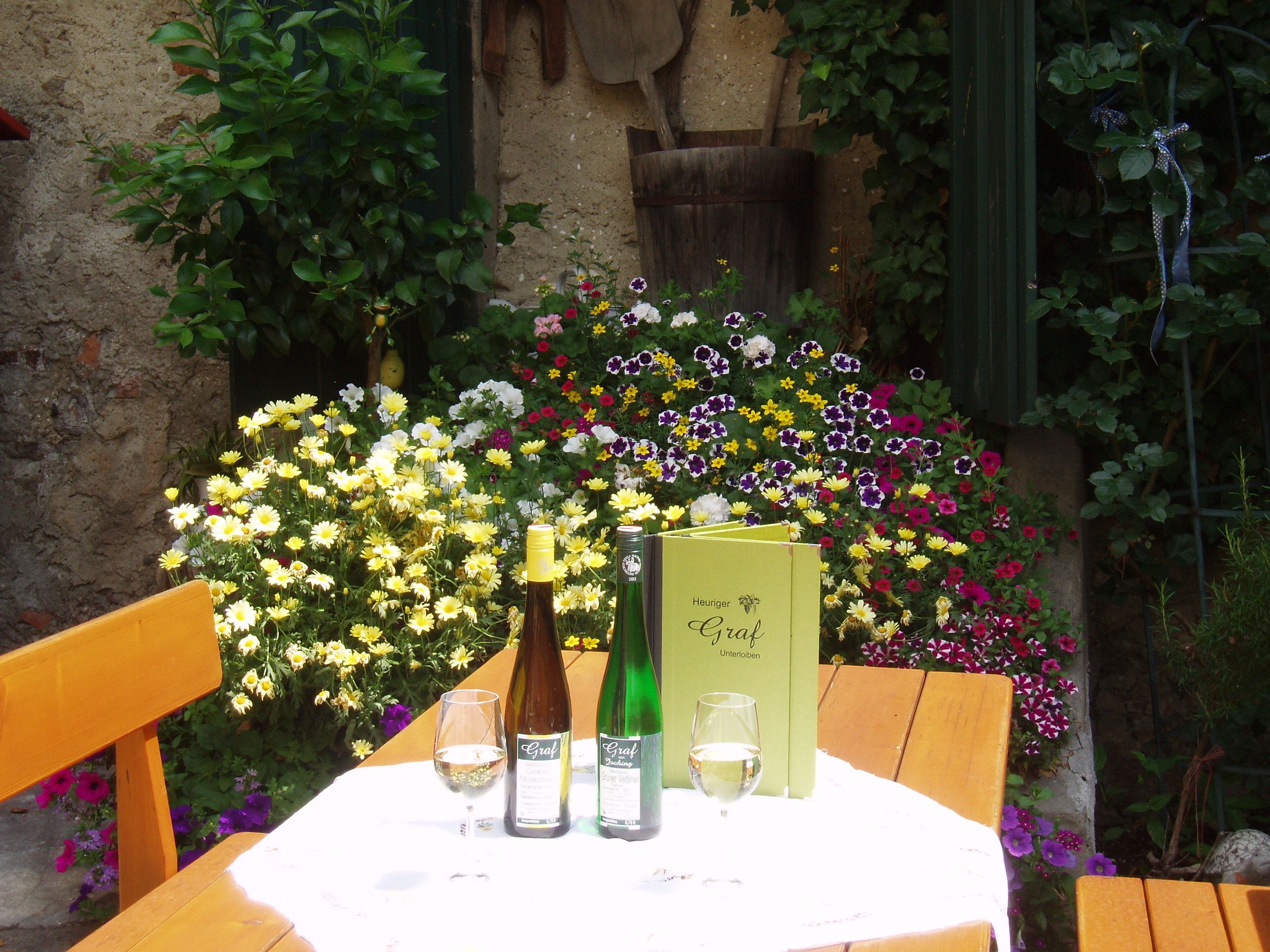 An outdoor table with bottles of wine, glasses and a menu, surrounded by colorful flowers.