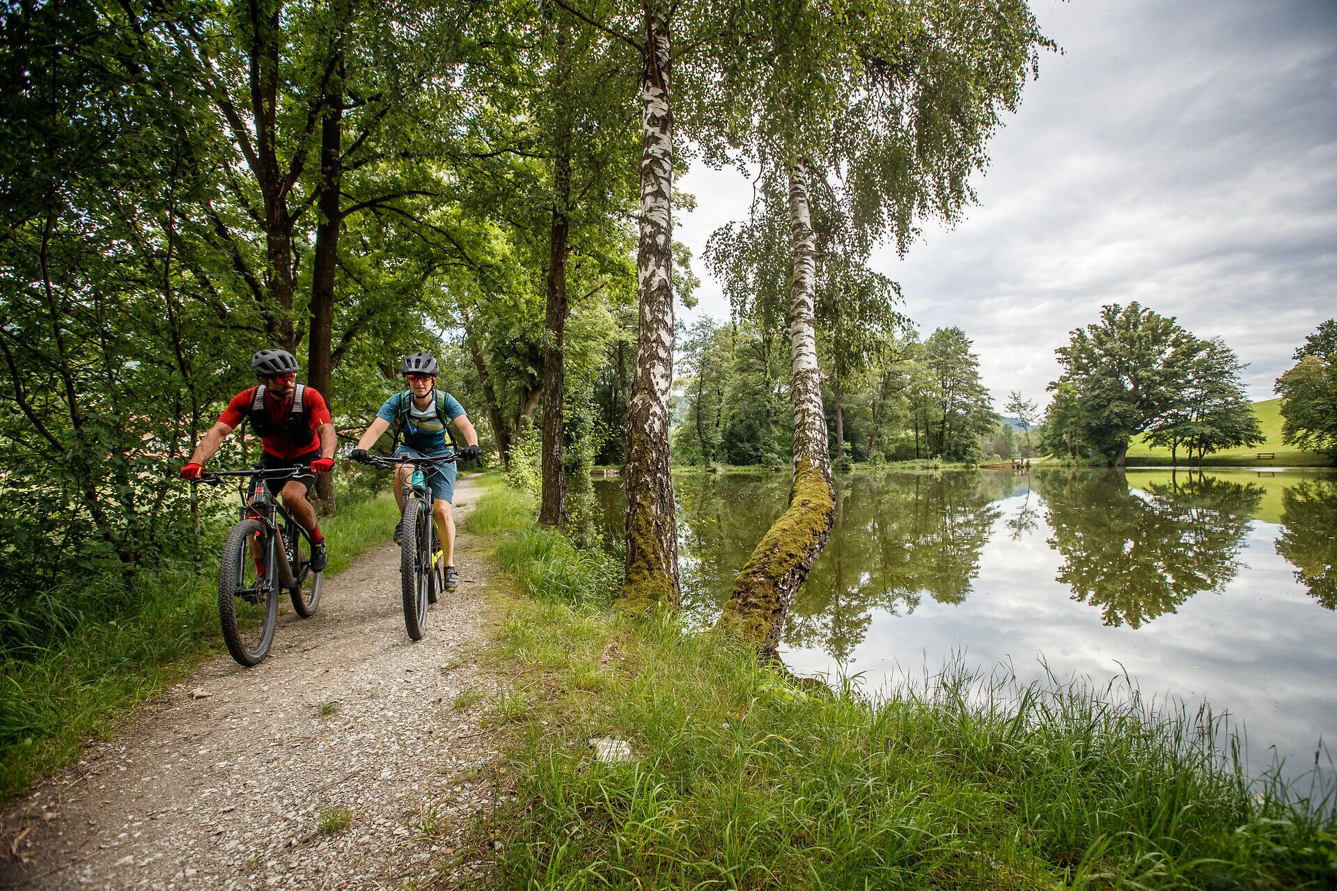 Two mountain bikers enjoy the fresh air and picturesque scenery along a quiet path lined with tall trees. The glistening lake reflects the clouds and the lush green of the surroundings, while the two cyclists immerse themselves in nature with joy and energy.