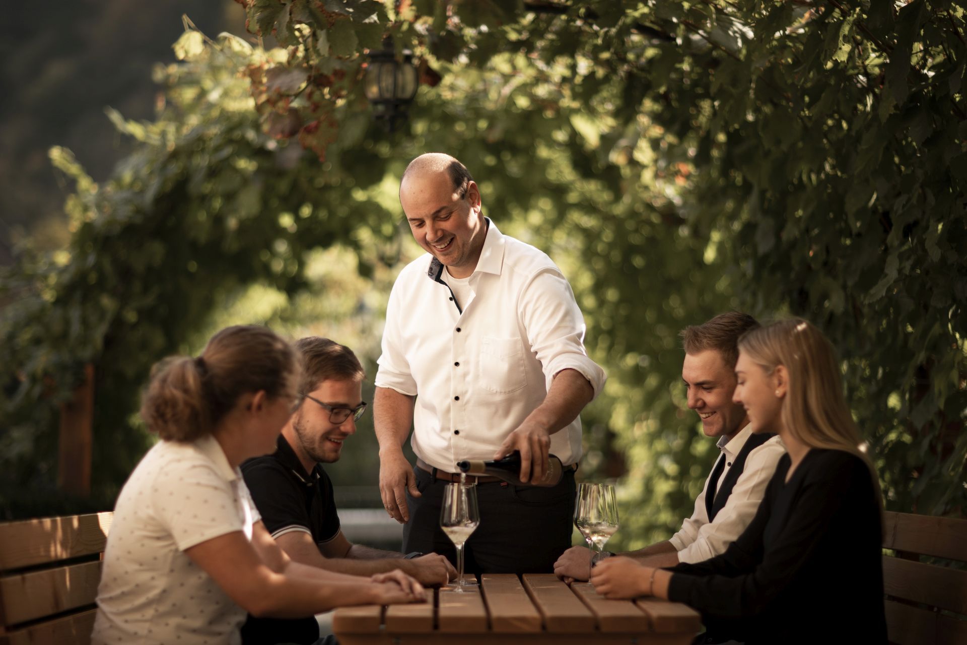 A man pours wine for four guests at an outdoor table surrounded by green vines.