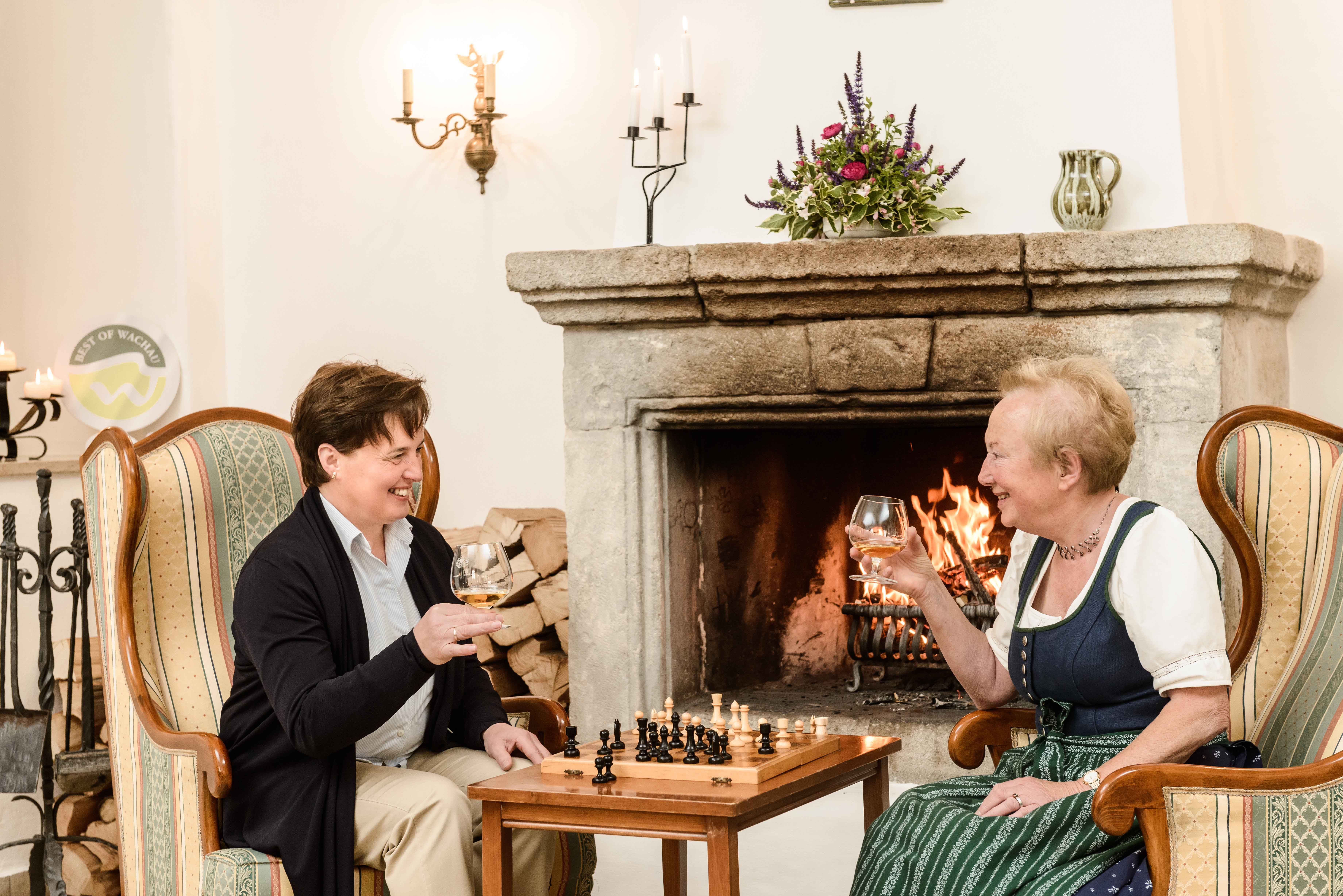 Two women are sitting in armchairs in front of a fireplace, drinking wine and playing chess.