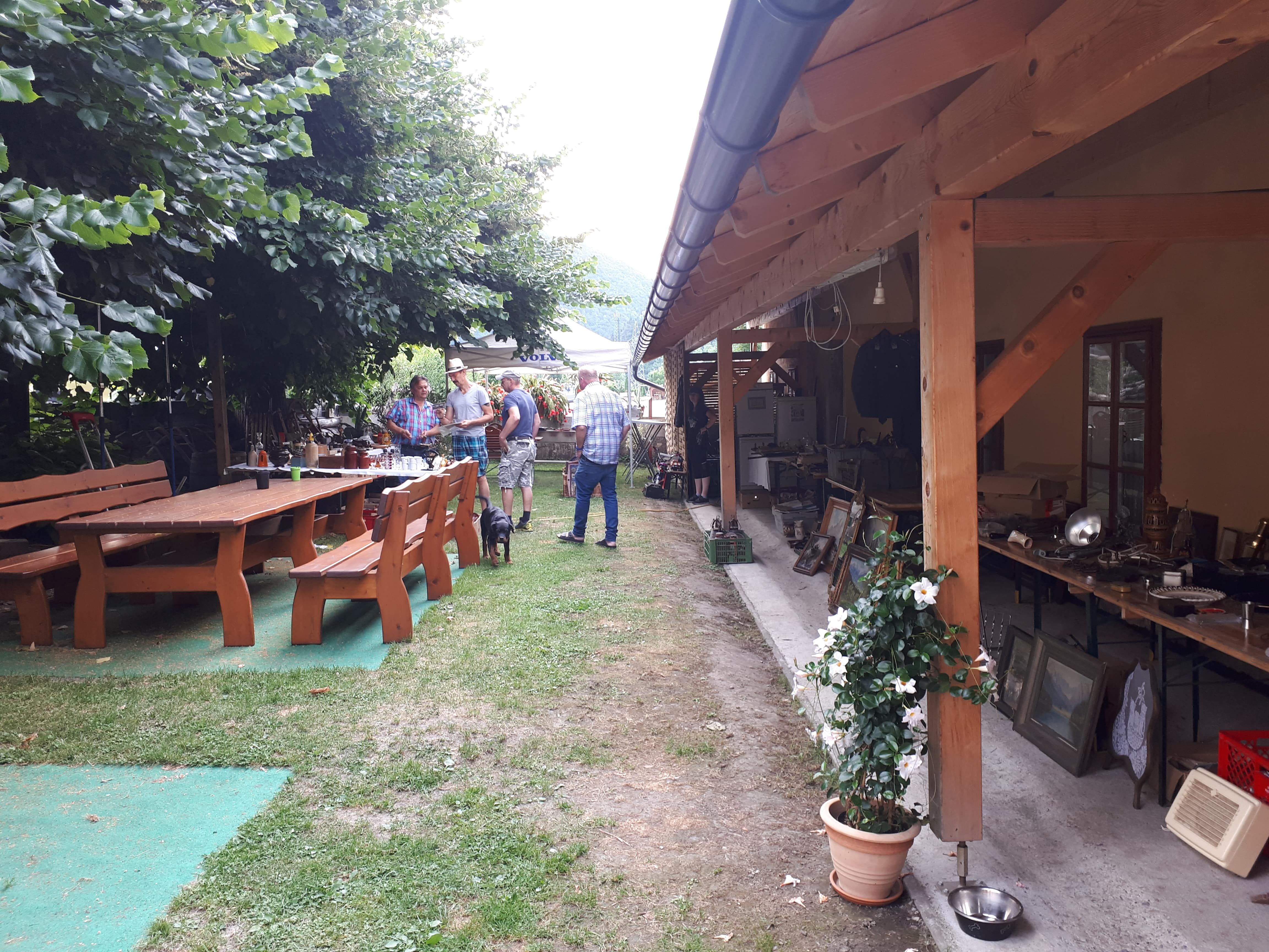 A garden with wooden tables and benches, people chatting, a dog, and a covered area with a flea market.