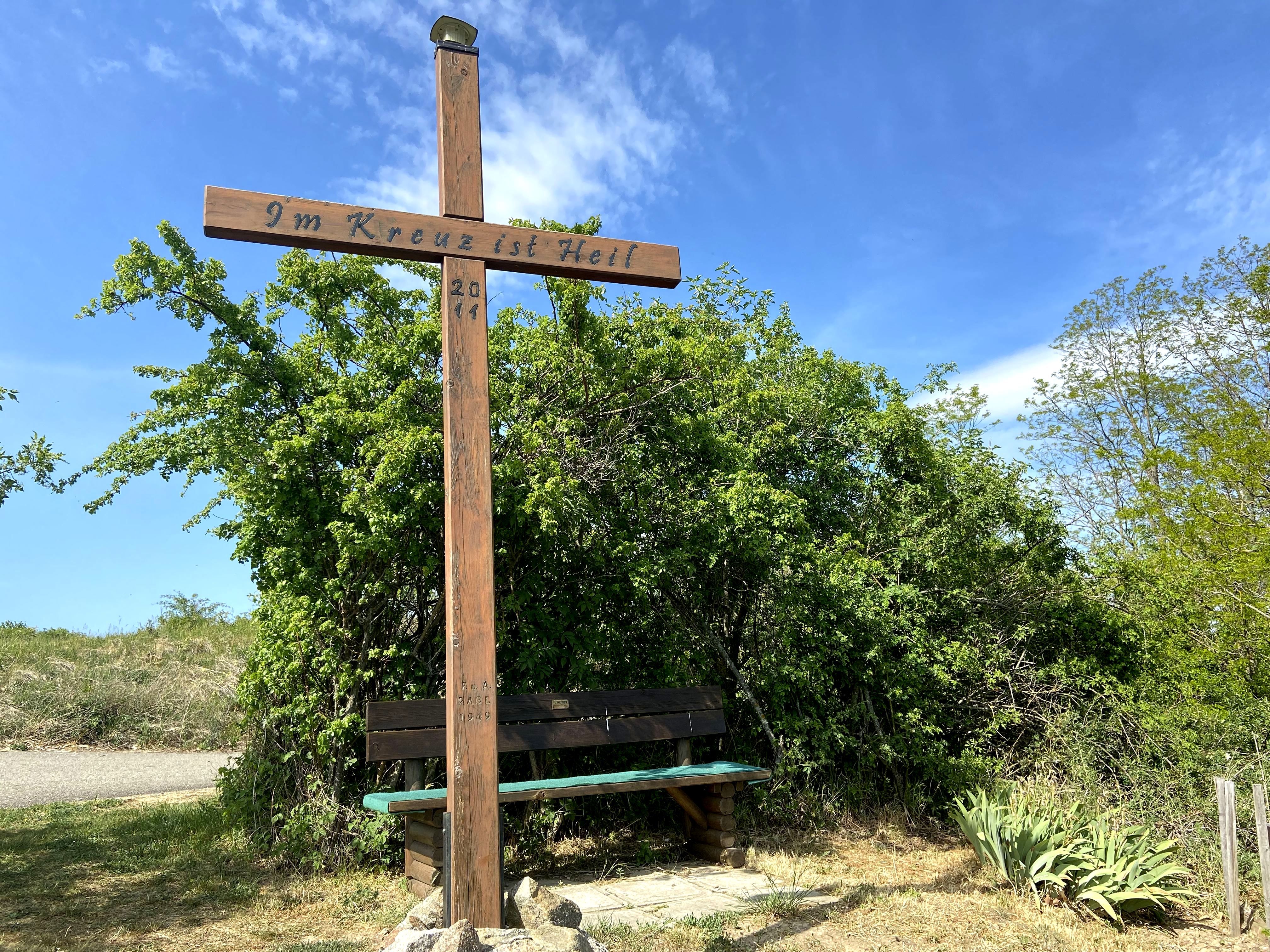 Wooden cross with inscription 'In the cross is salvation' and bench, surrounded by bushes and blue sky.