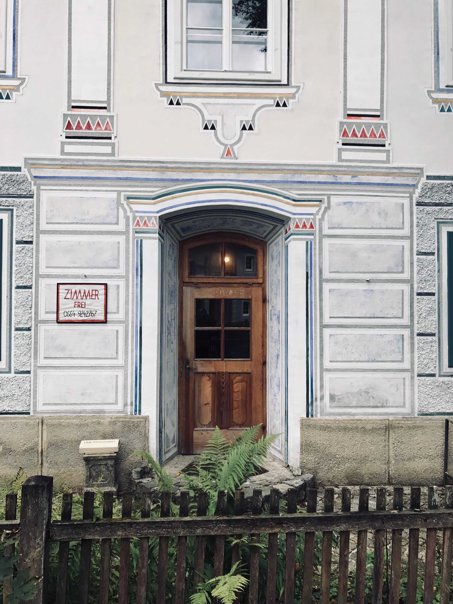 Entrance to an ornate building with a wooden door and decorative paintings, surrounded by a wooden fence and ferns.