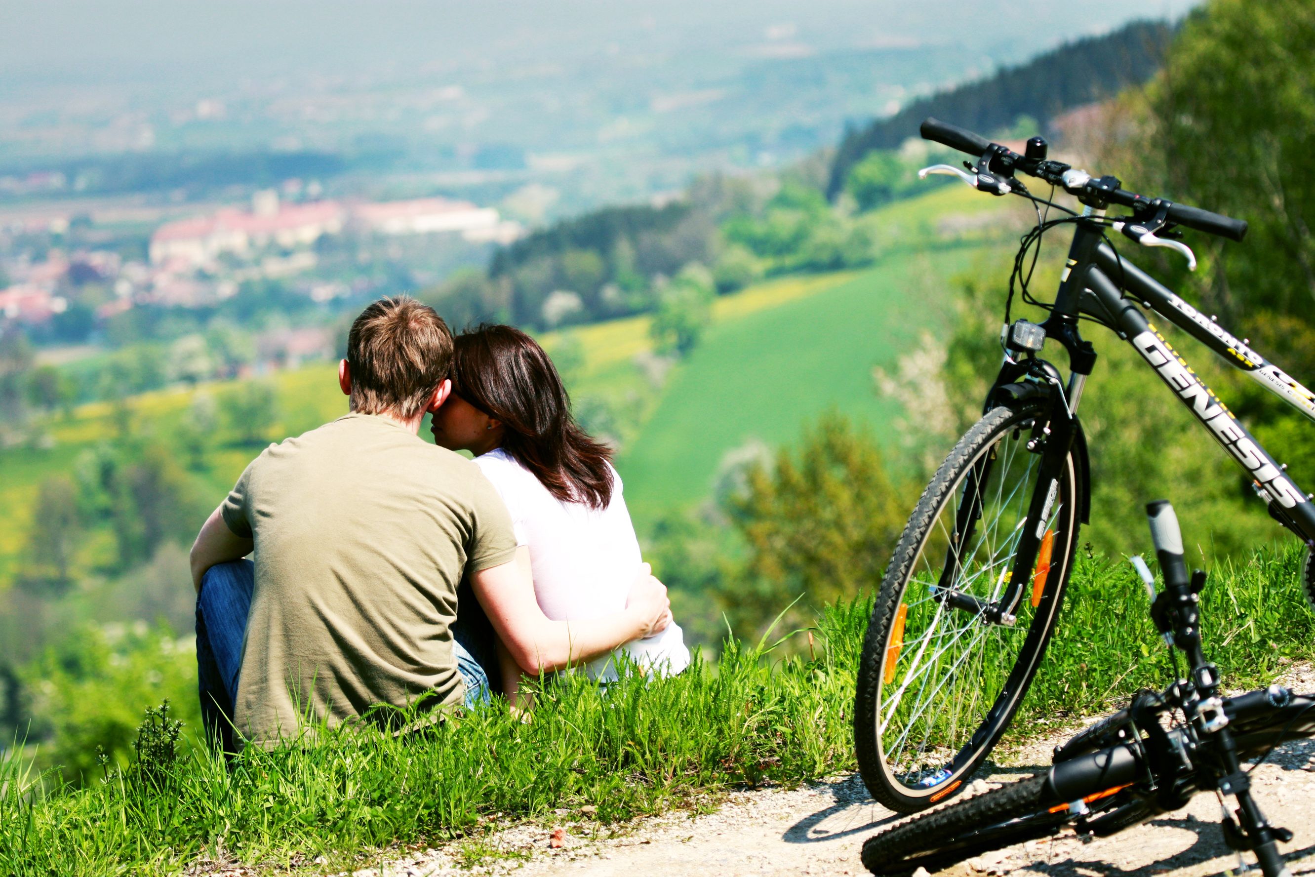 A couple is sitting on a meadow with a view of the landscape, with bicycles parked next to them.