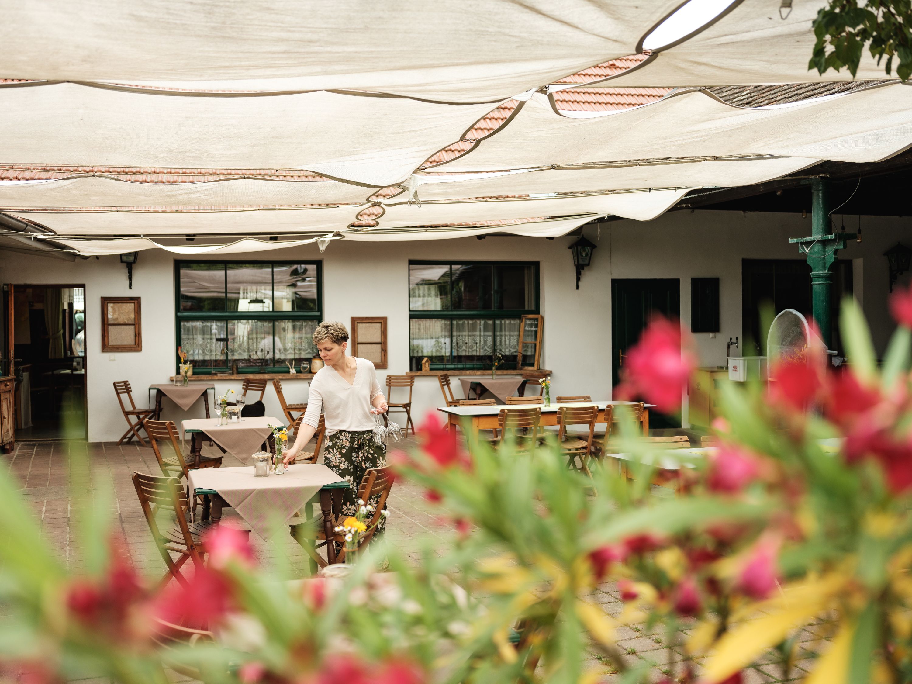 A woman prepares tables in a covered outdoor area of an inn. Blurred flowers can be seen in the foreground.