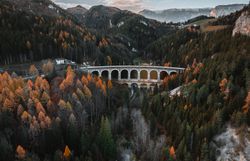 Die majestätische Semmeringbahn schlängelt sich durch die bunten Herbstwälder der Wiener Alpen. Goldene und rote Blätter umrahmen die beeindruckenden Bögen der Brücke, während die sanften Hügel im Hintergrund eine malerische Kulisse bieten. Ein Ort, der die Seele berührt und zum Verweilen einlädt.