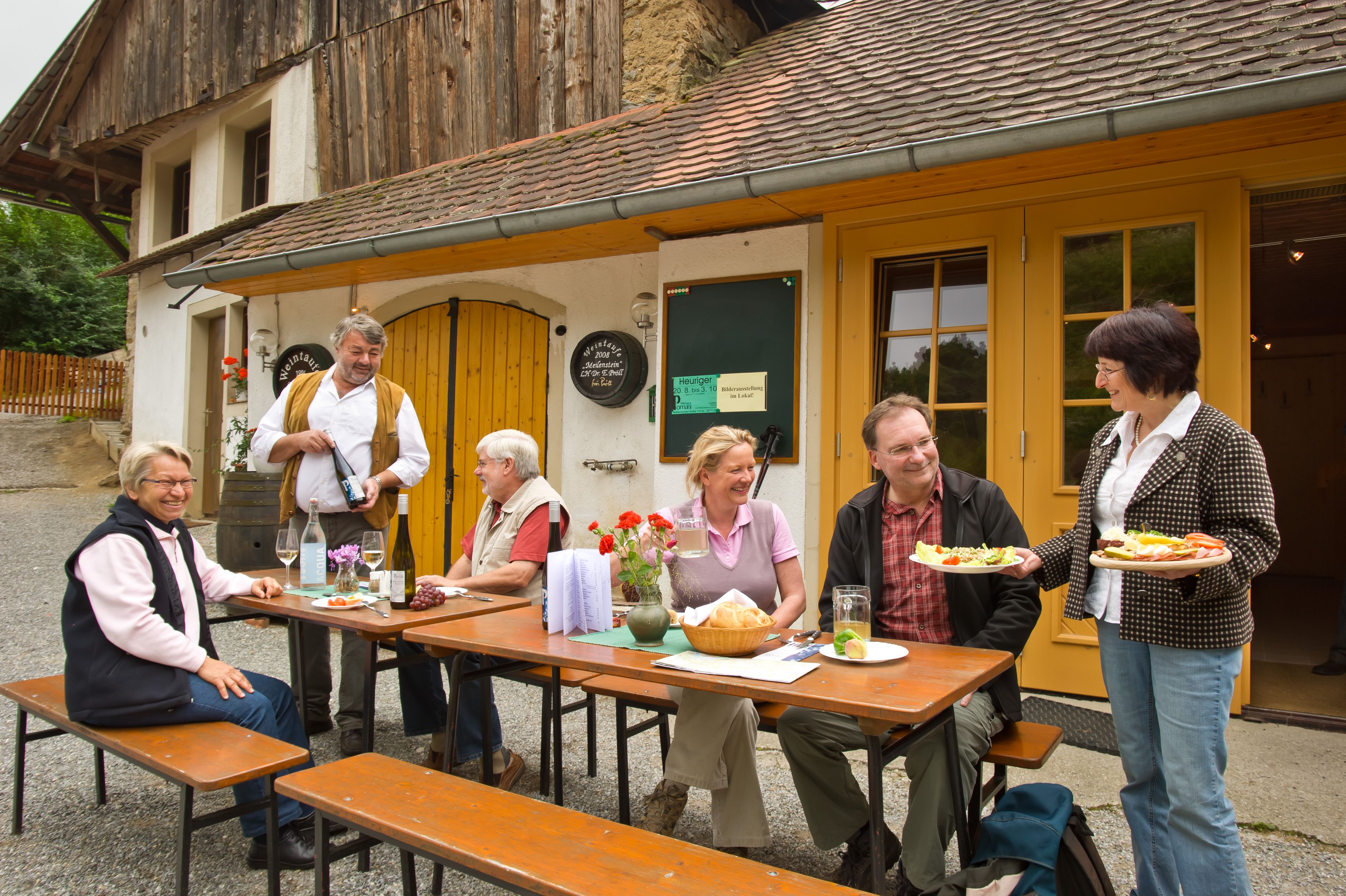 Group of people sitting at a table in front of a rustic building enjoying an outdoor meal.