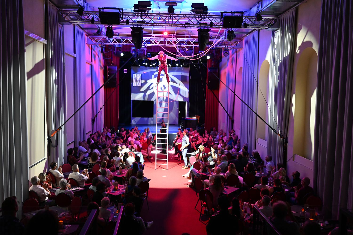 An artist balances on a ladder in a crowded theater with colorful lighting.