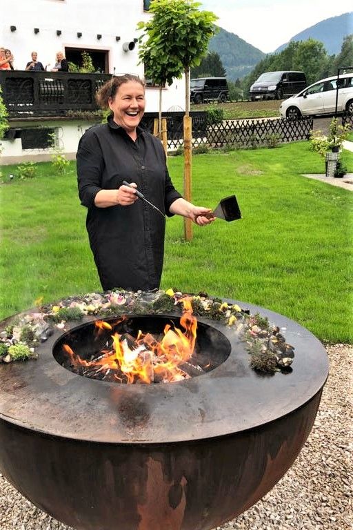 A woman stands laughing by an outdoor fireplace, surrounded by a green landscape and a building in the background.