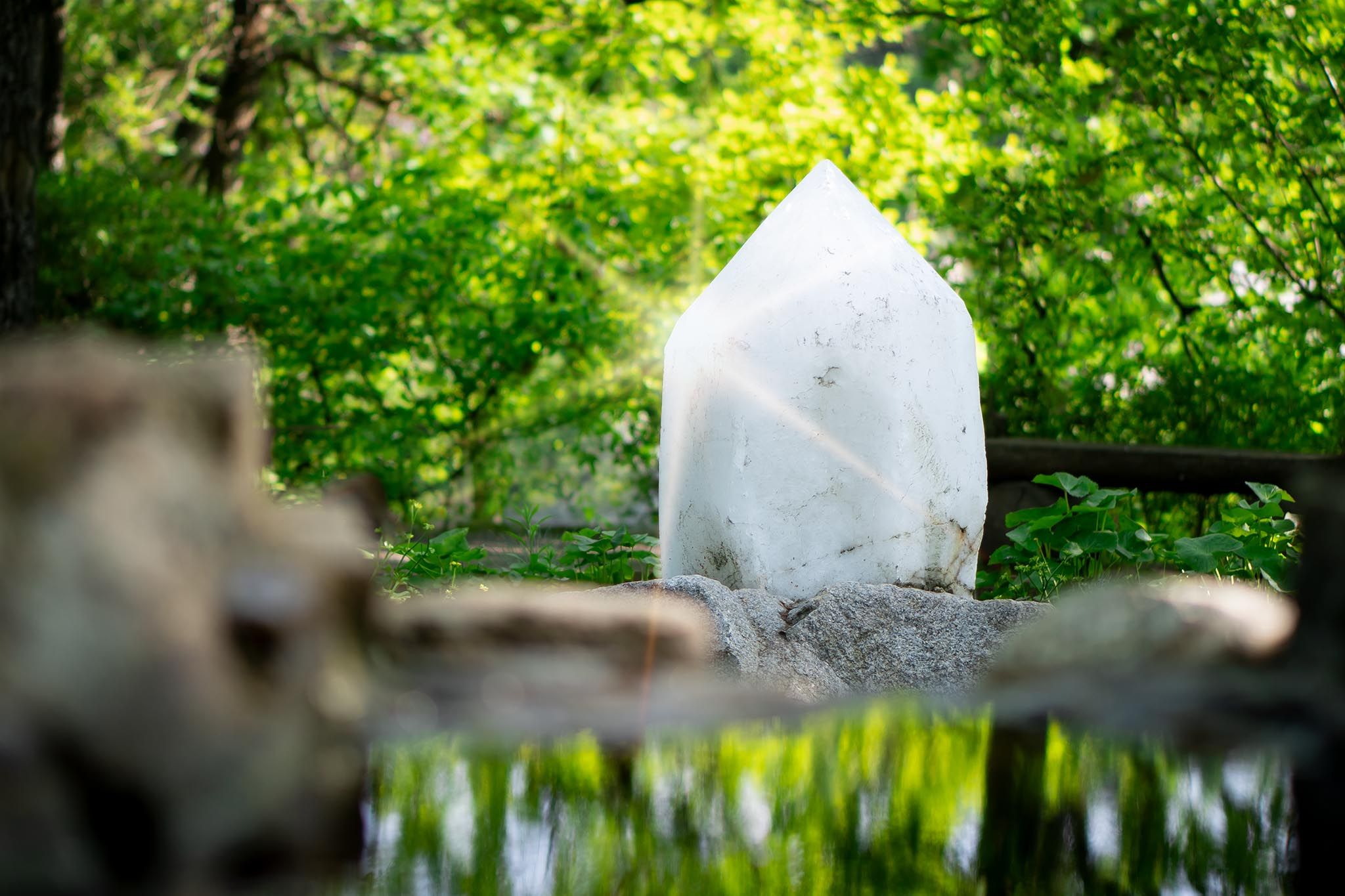 A large white crystal stands in a green forest, surrounded by plants and reflected in a small pond.