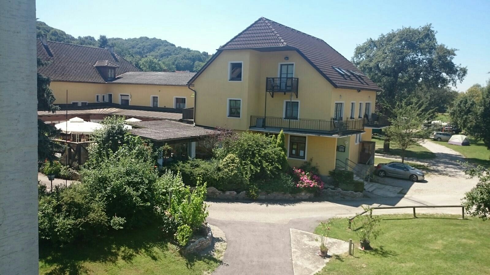 Yellow building with terrace and garden, surrounded by trees and a car in the parking lot.