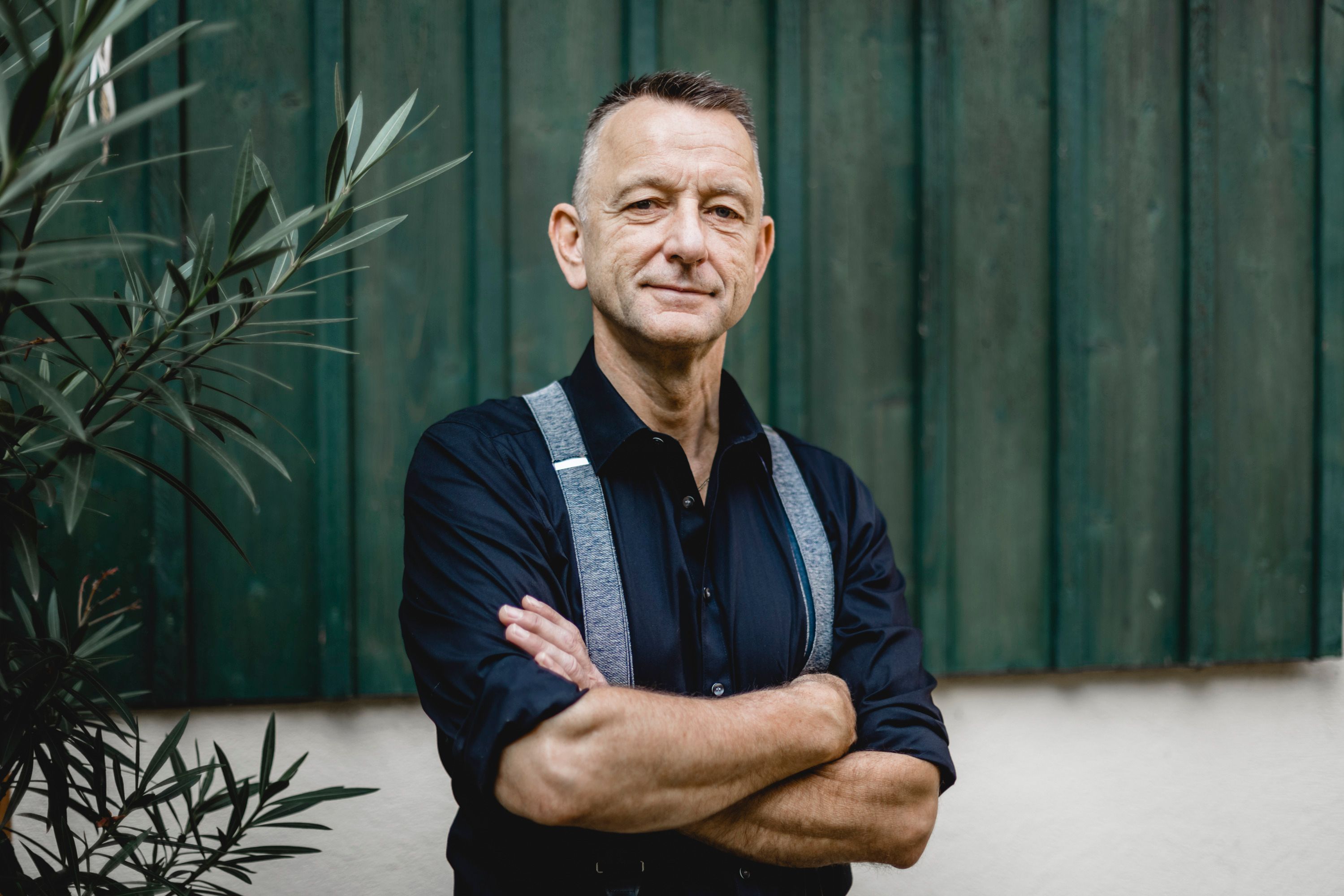 A man in a dark shirt and suspenders stands with his arms folded in front of a green wooden wall.