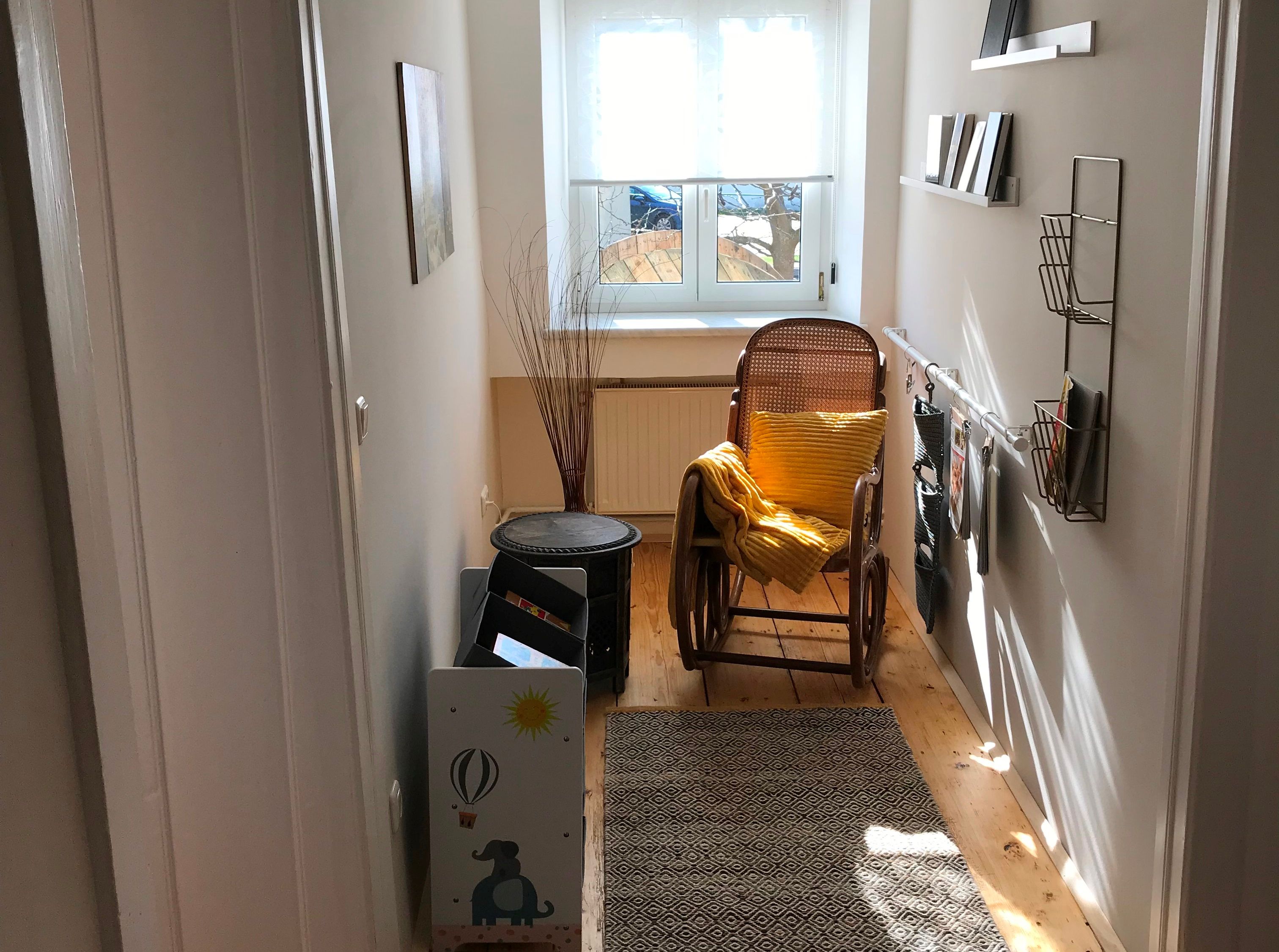 A narrow entrance area with a rocking chair, yellow cushions, carpet and shelves against the wall. A window at the end of the hallway.