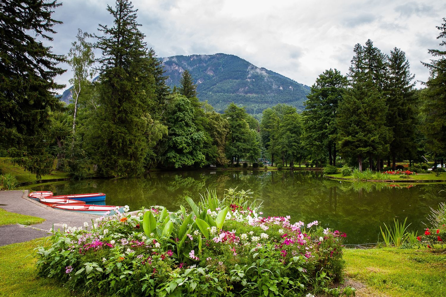 A picturesque park with a pond, surrounded by trees and flowers, with mountains in the background.