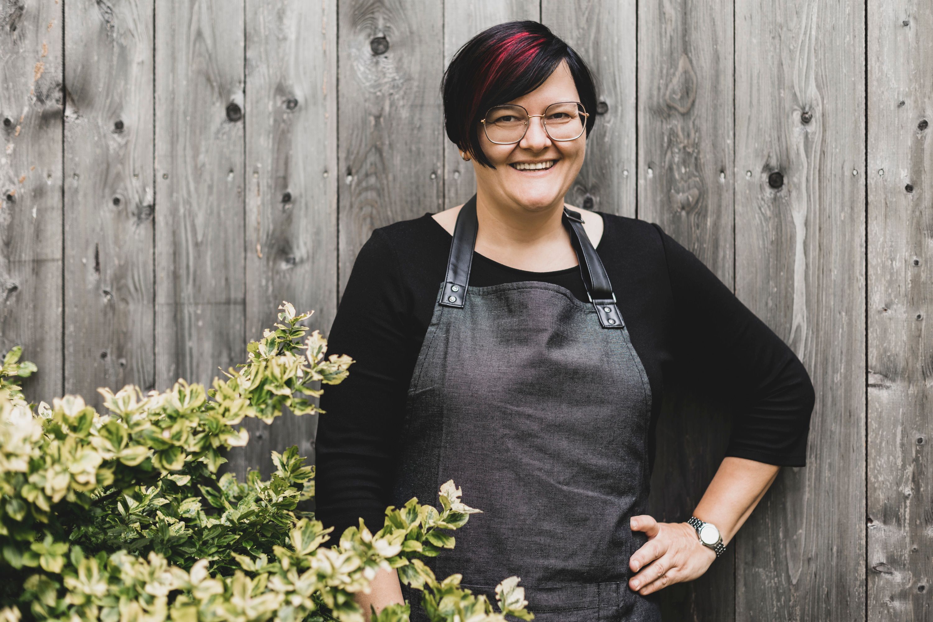 A woman with glasses and an apron stands smiling in front of a wooden wall next to a bush.