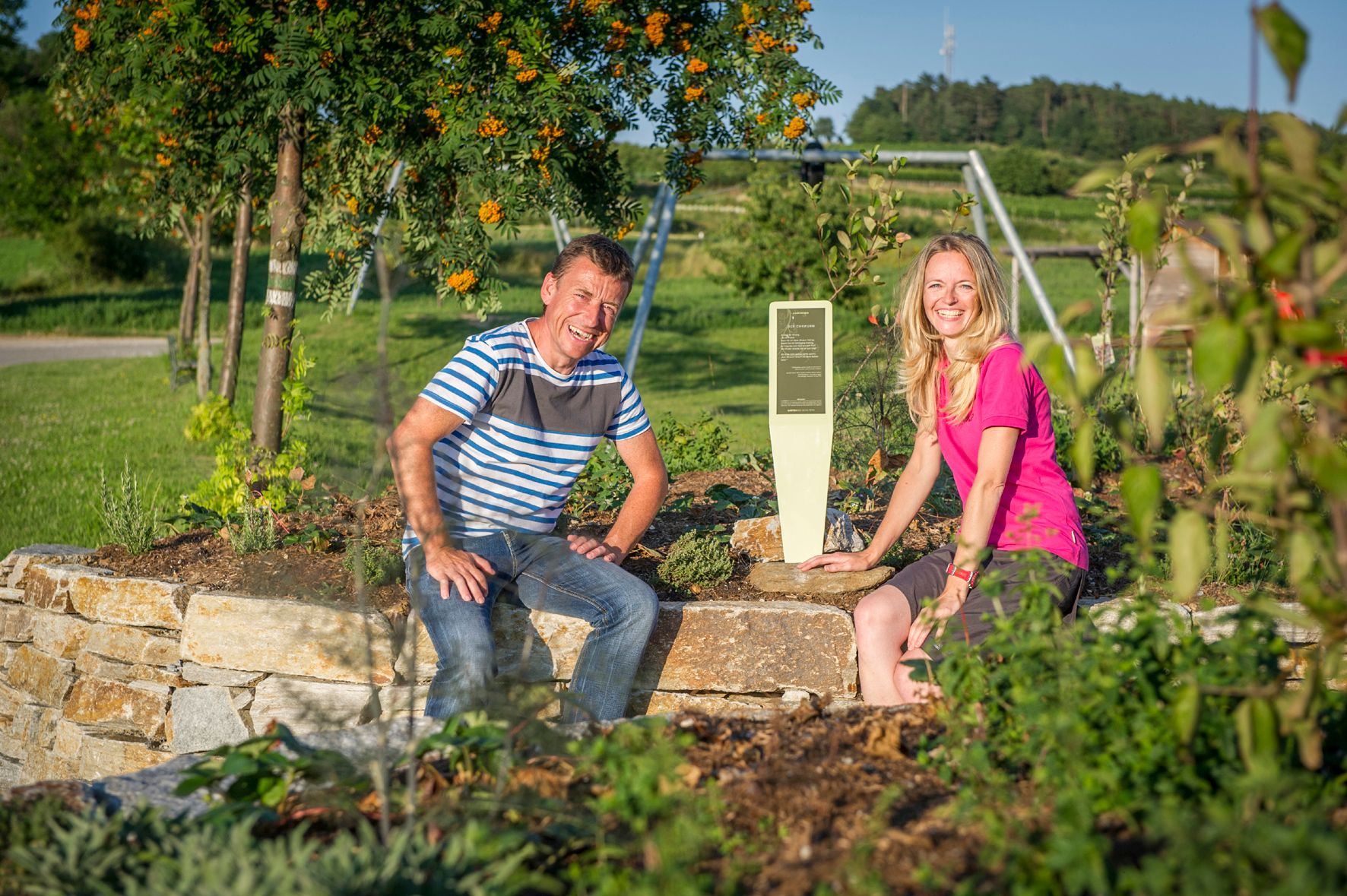 Two people are sitting on a stone wall in a garden with plants and a sign.