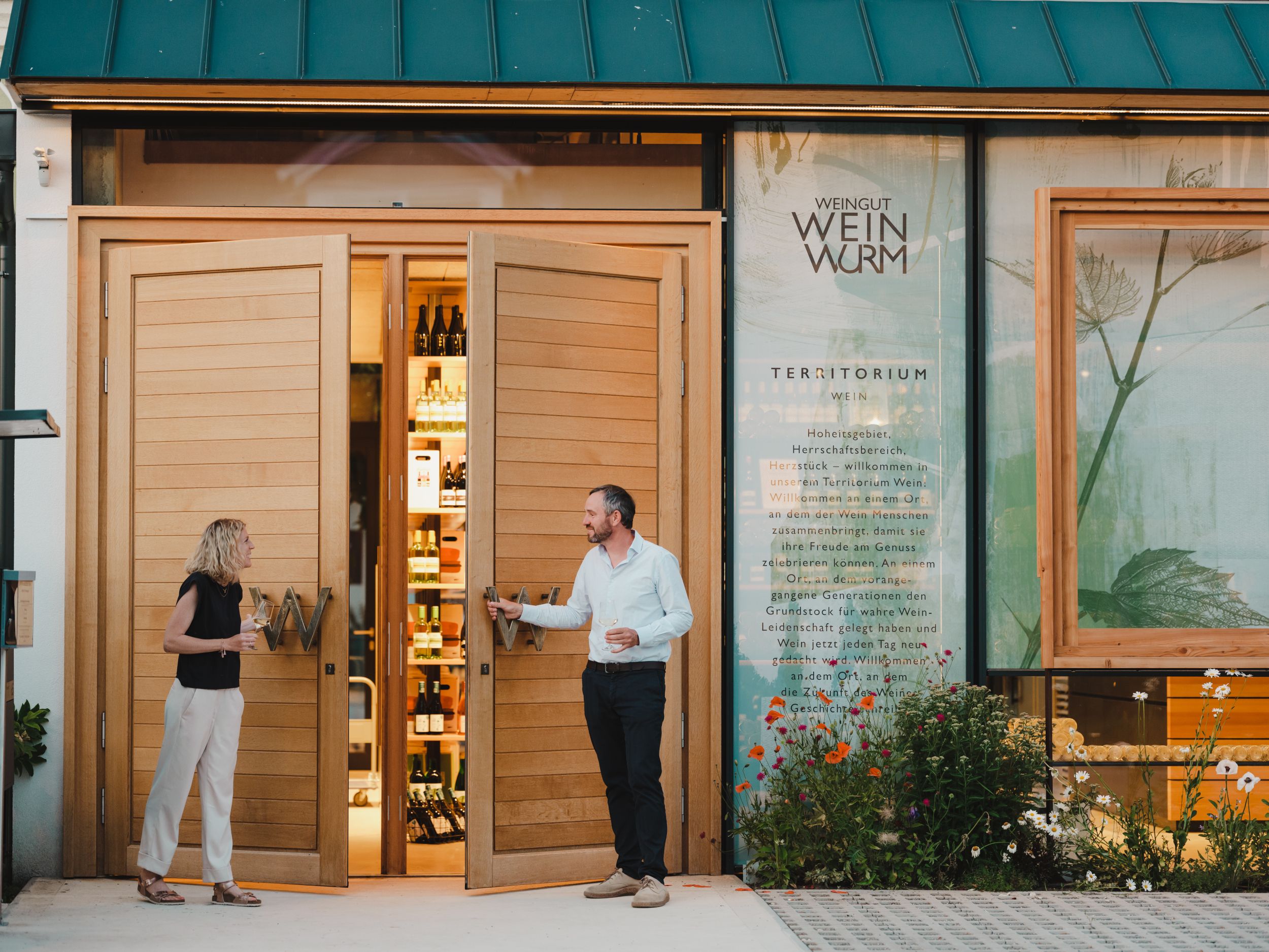 Two people are standing in front of a winery with open wooden doors showing bottles inside.