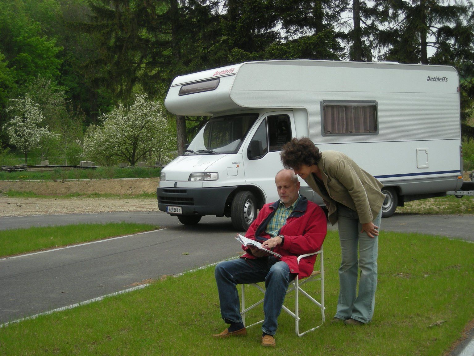A man sits on a camping chair in front of a camper van while a woman looks over his shoulder. Trees and a meadow can be seen in the background.