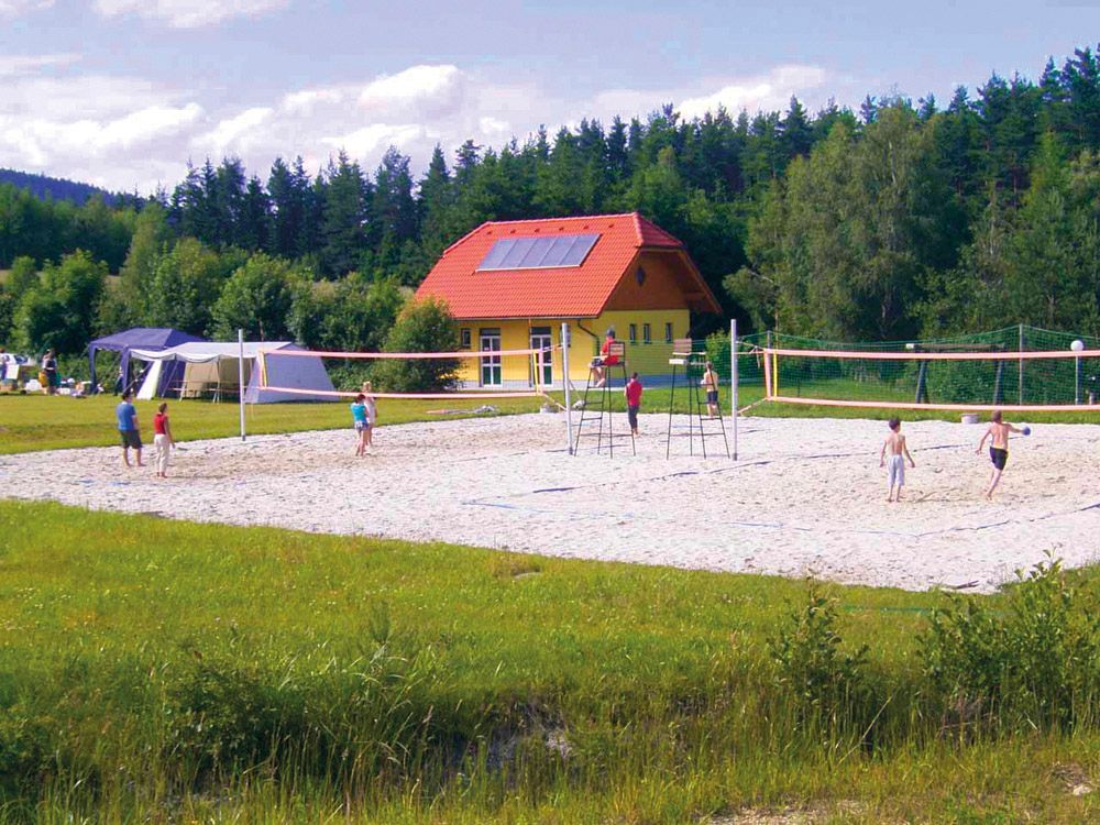 People play volleyball on a sand court in front of a yellow building with a red roof, surrounded by trees.