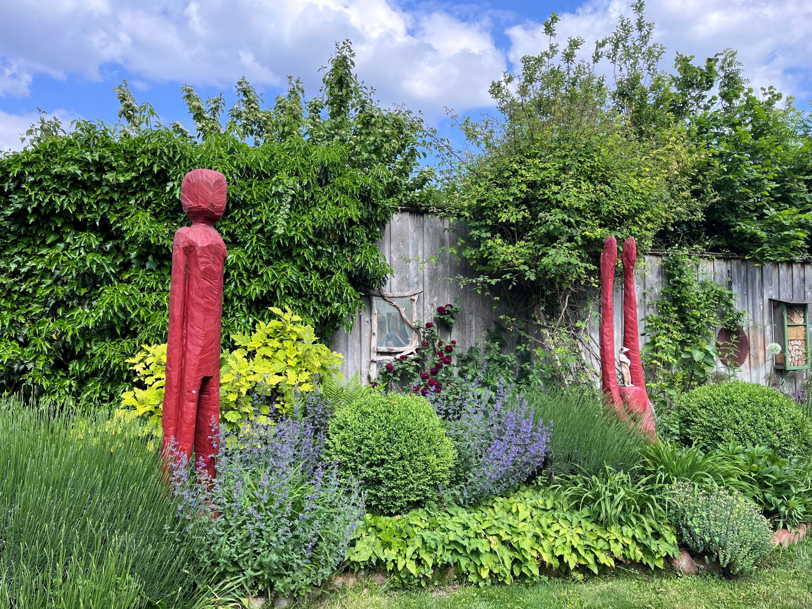 Garden with red sculptures, surrounded by green plants and a wooden wall in the background.