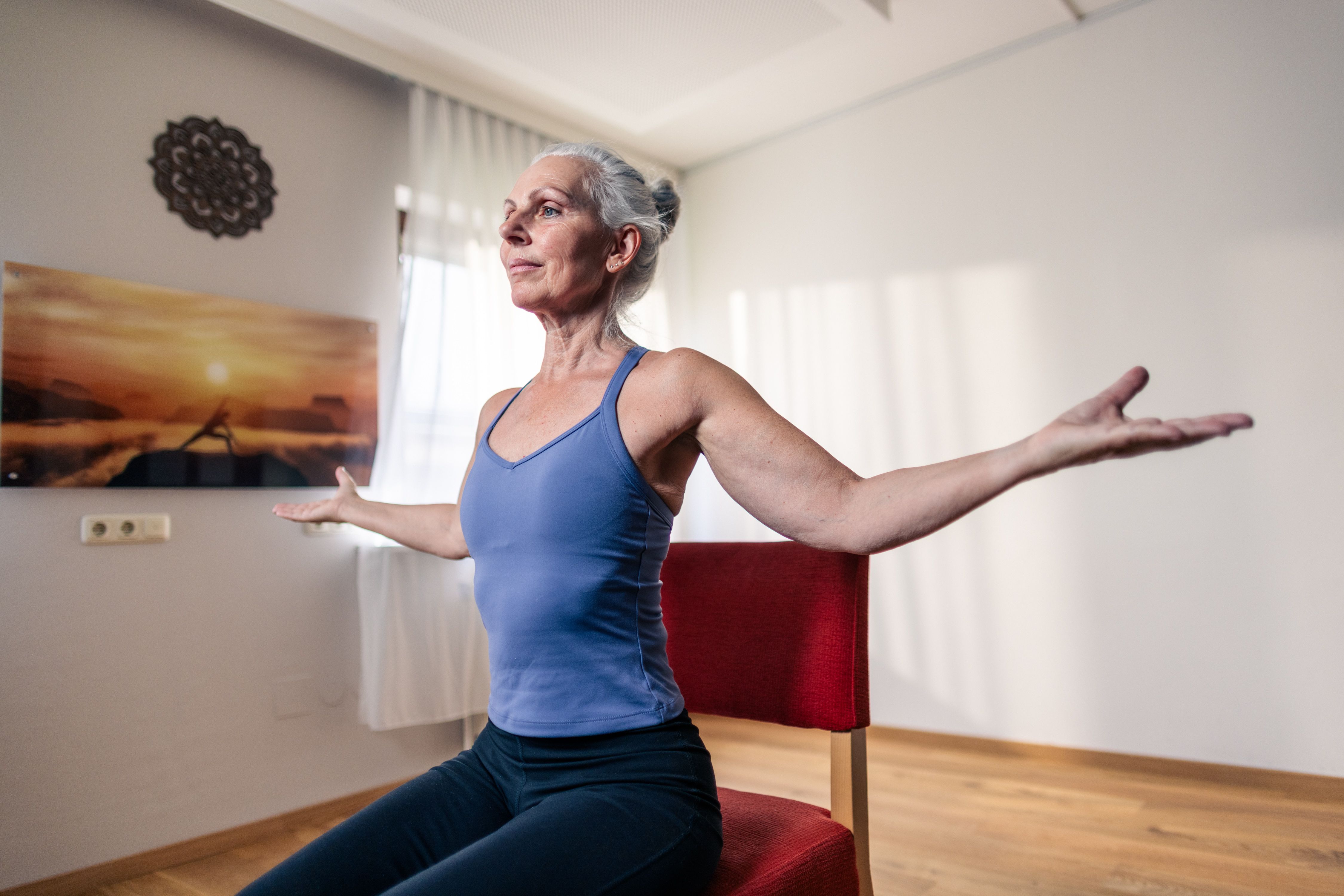 An elderly woman is doing yoga on a chair in a brightly lit room.