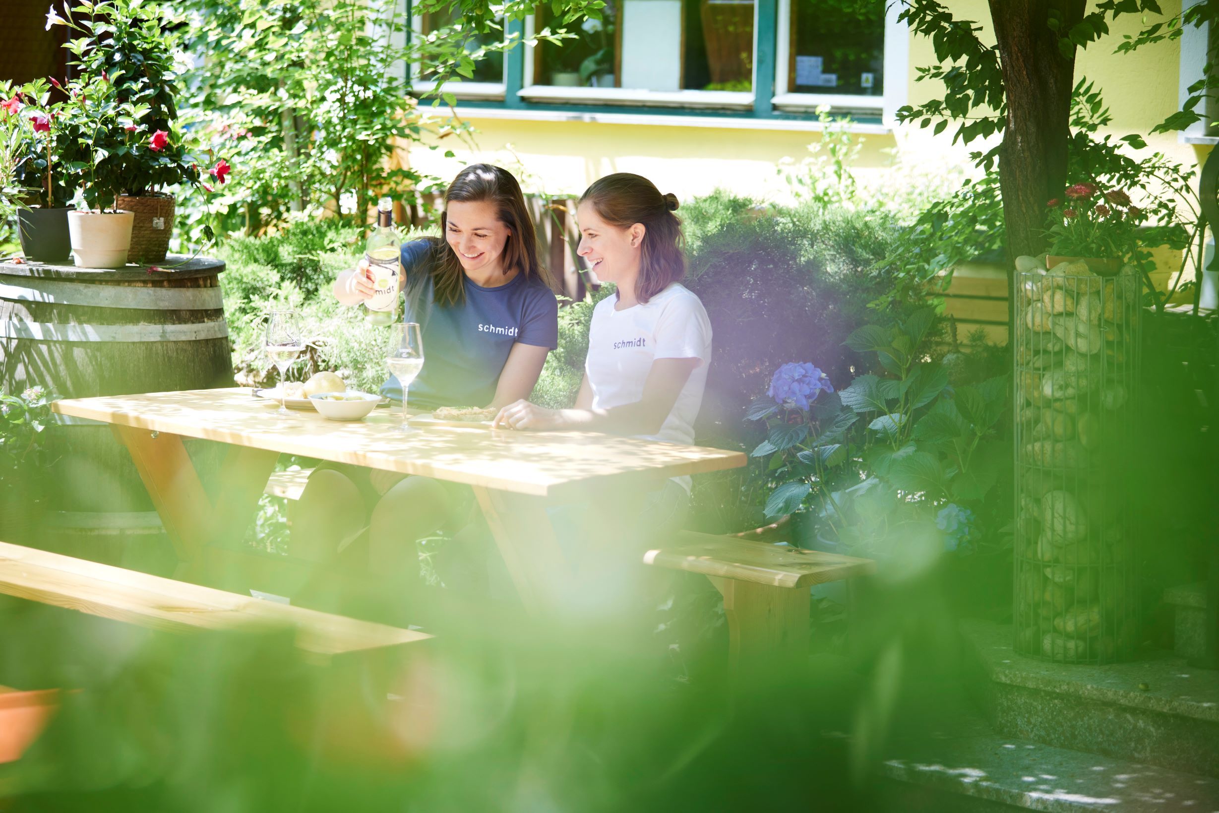 Two women sit at a wooden table in the garden and enjoy some wine.